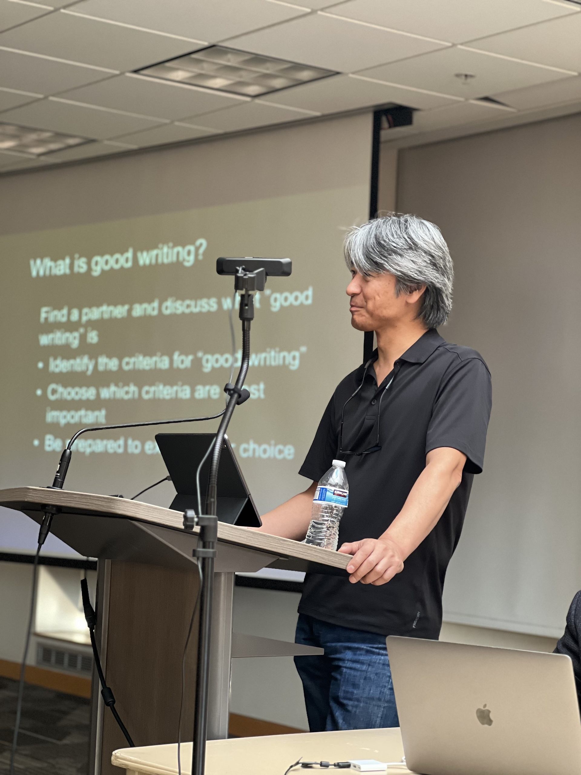 A photo of Paul Kei Matsuda delivering his presentation. On the background, the slide includes the following text: [What is good writing?​ Find a partner and discuss what ”good writing” is. ​Identify the criteria for “good writing”​. Choose which criteria are most important​. Be prepared to explain your choice​.]