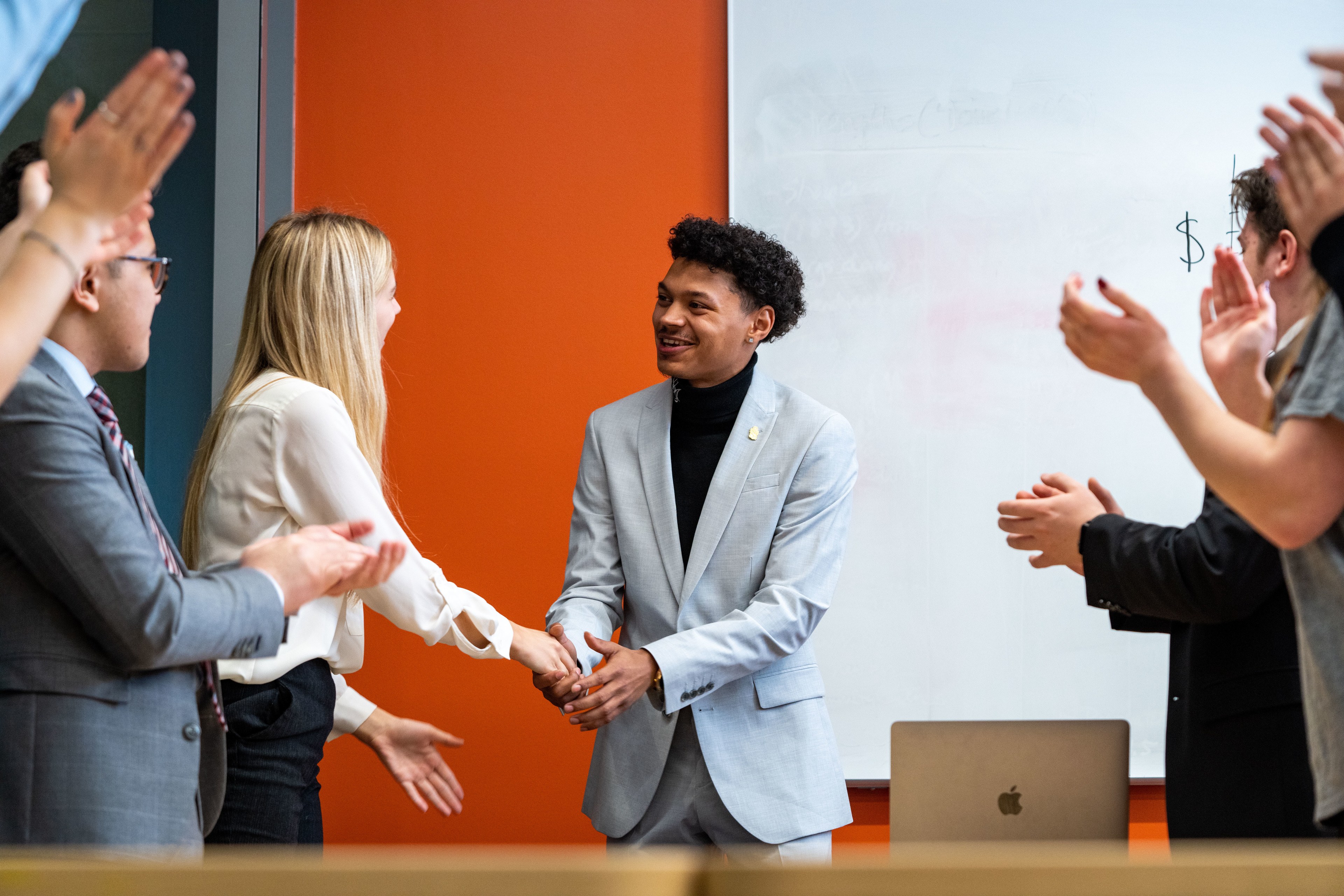 A business management student shakes hands with a classmate after a presentation while other students applaud.