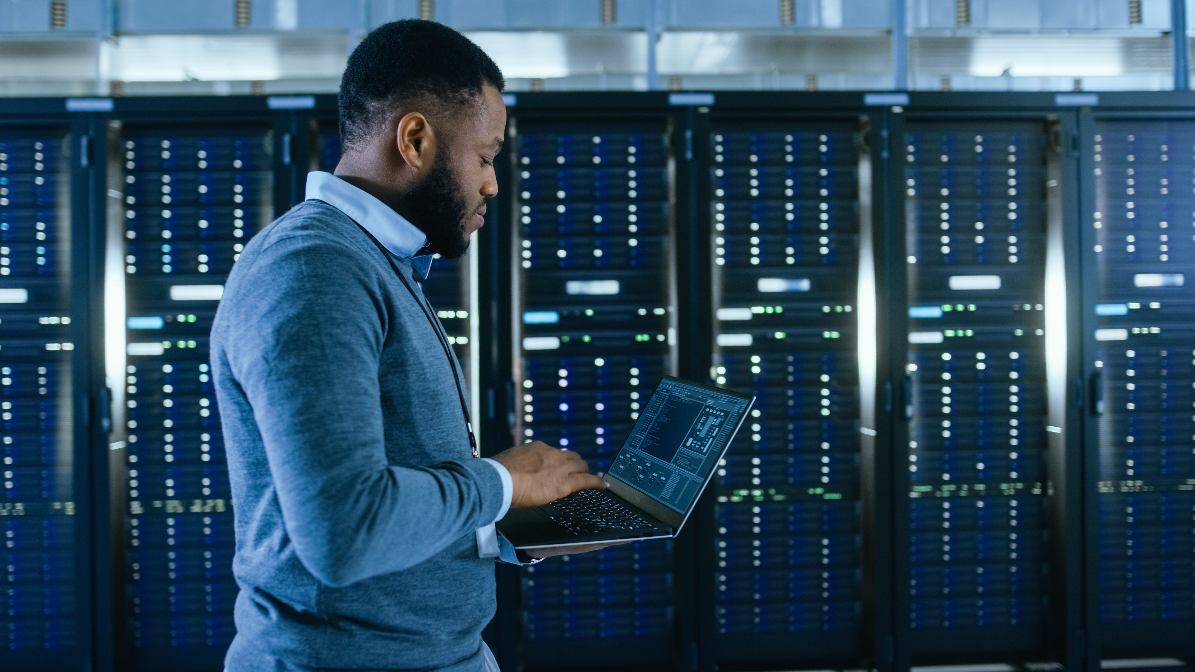 Black Data Center IT Technician Walking Through Server Rack Corridor with a Laptop Computer. He is Visually Inspecting Working Server Cabinets.