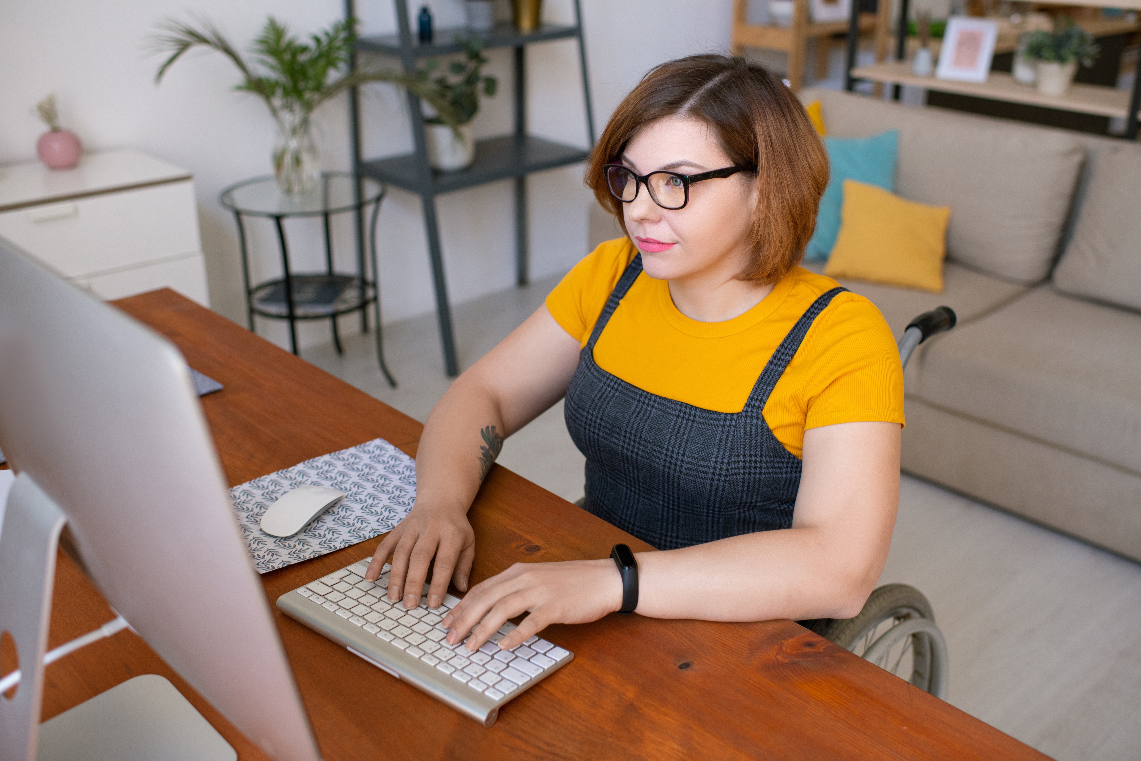 Young female in casualwear sitting in wheelchair by desk in front of computer