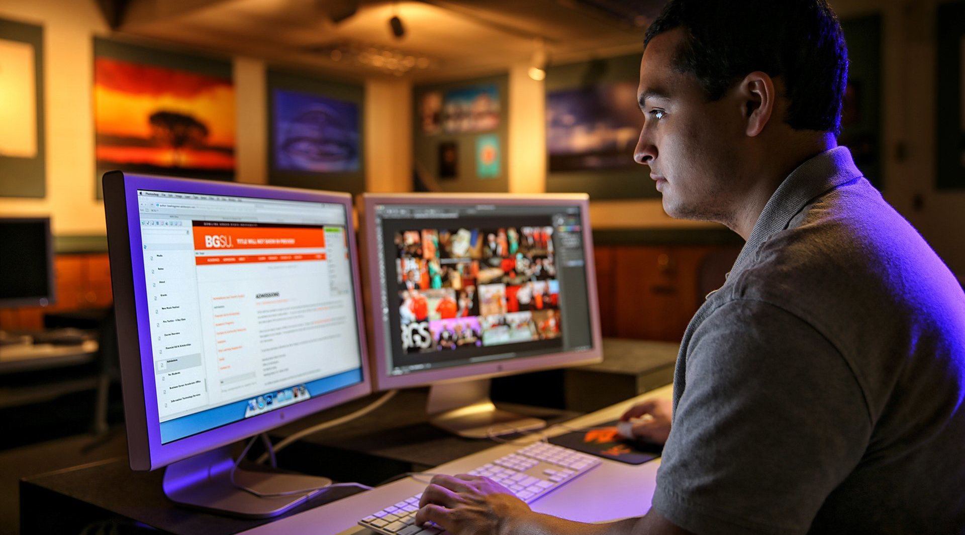 A male VCT student at BGSU sits in front of two computer monitors and works on the BGSU website.