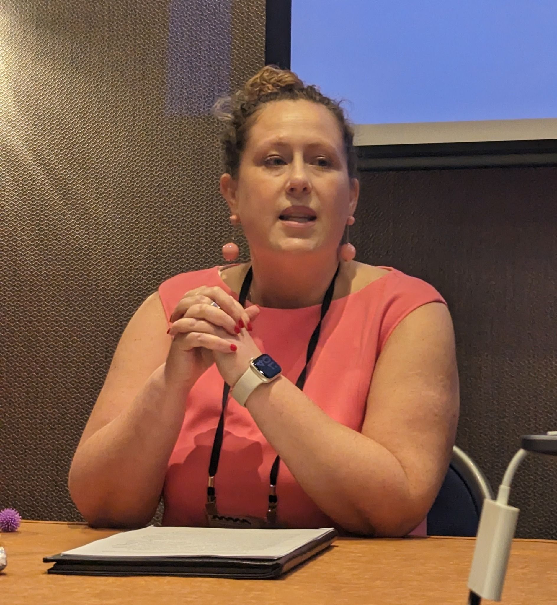 White woman in pink dress is seated at table, talking to audience 