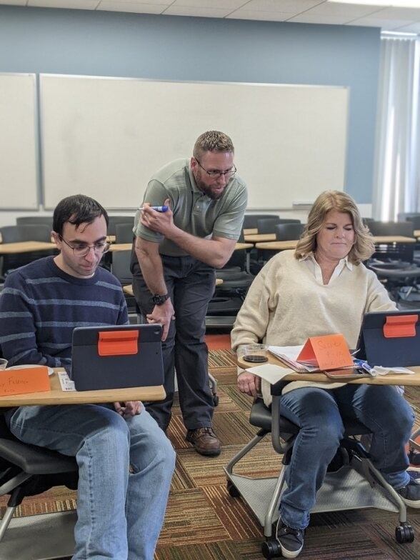 Two people sit at their computers with a third person assisting them 