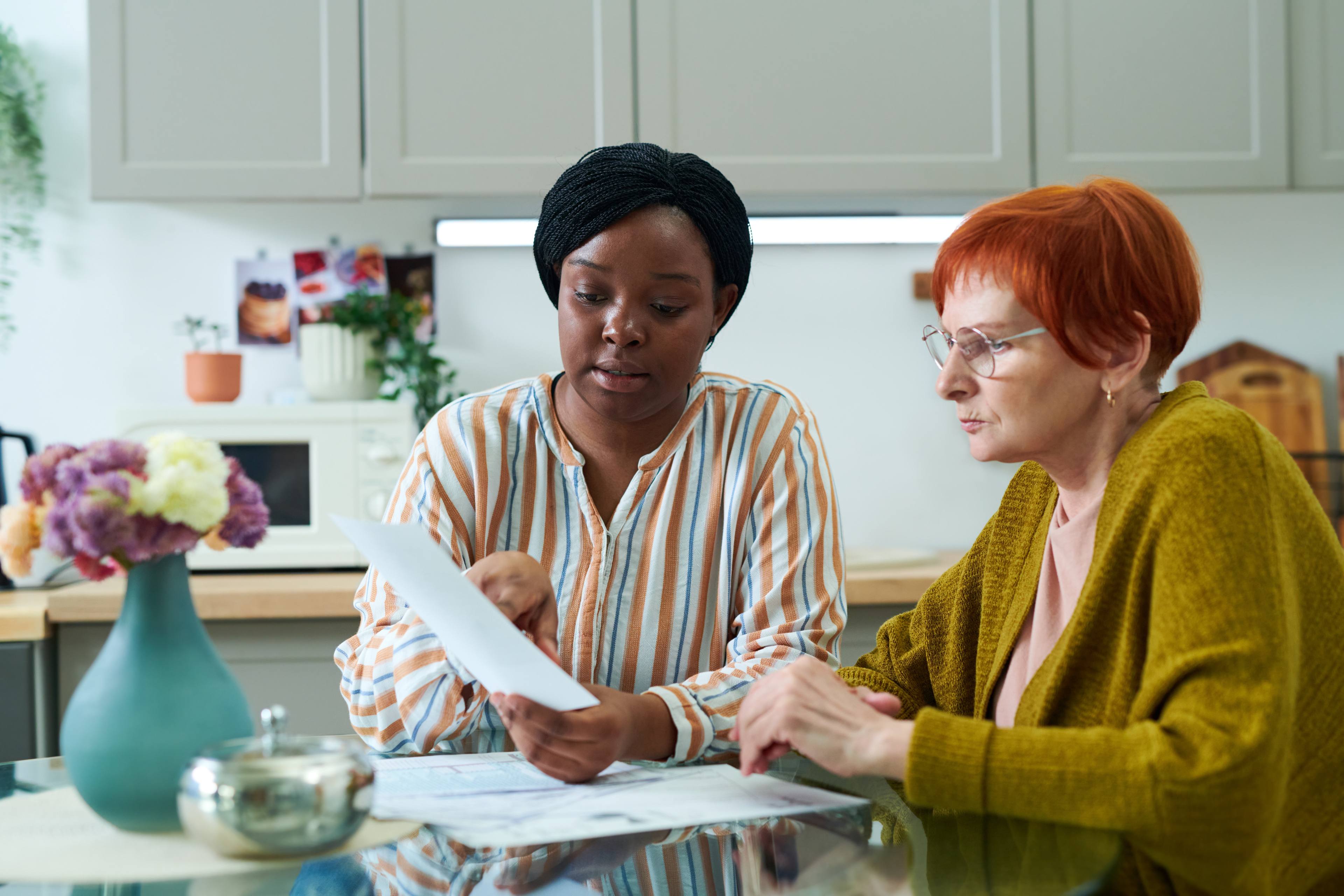 African young volunteer sitting at kitchen table together with senior woman and helping with bills