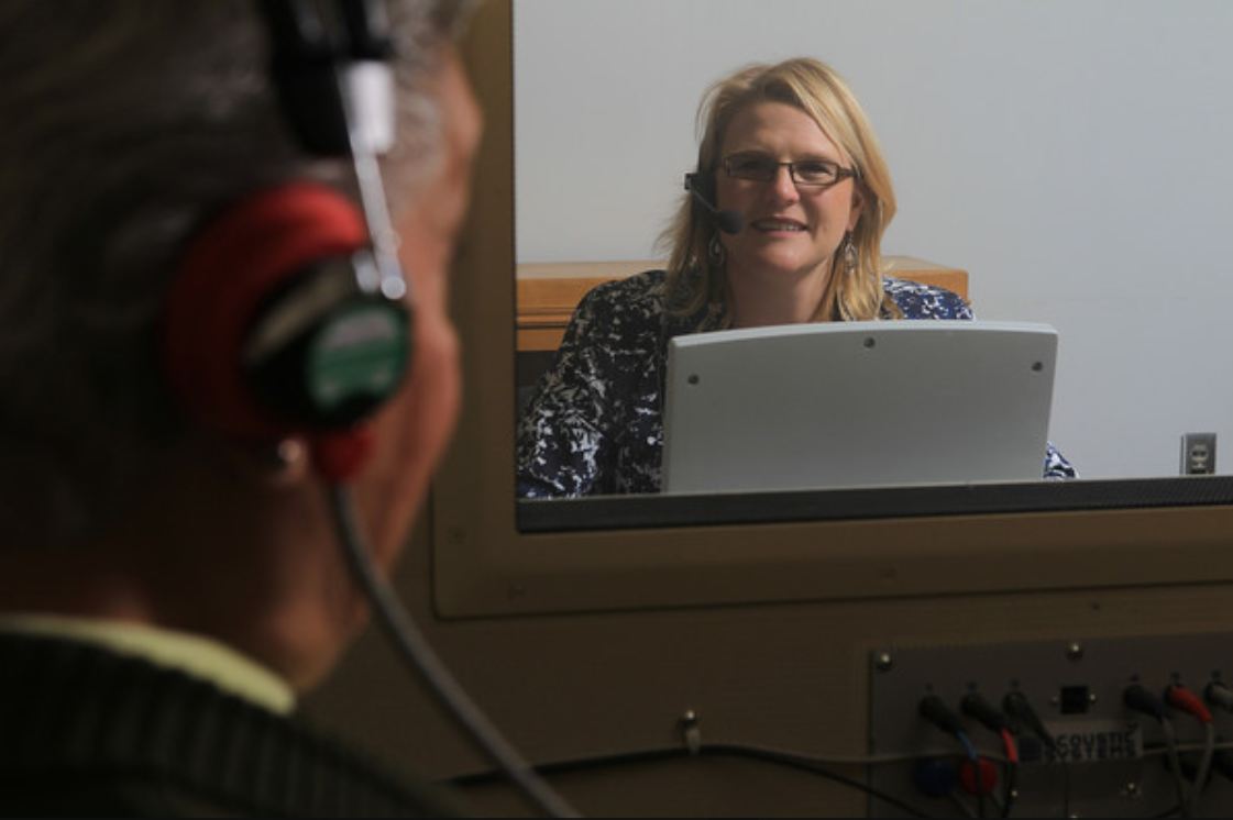 A man sits inside a booth with headphones on. He is having his hearing tested. A smiling audiologist sits outside the booth, visible through a glass window.