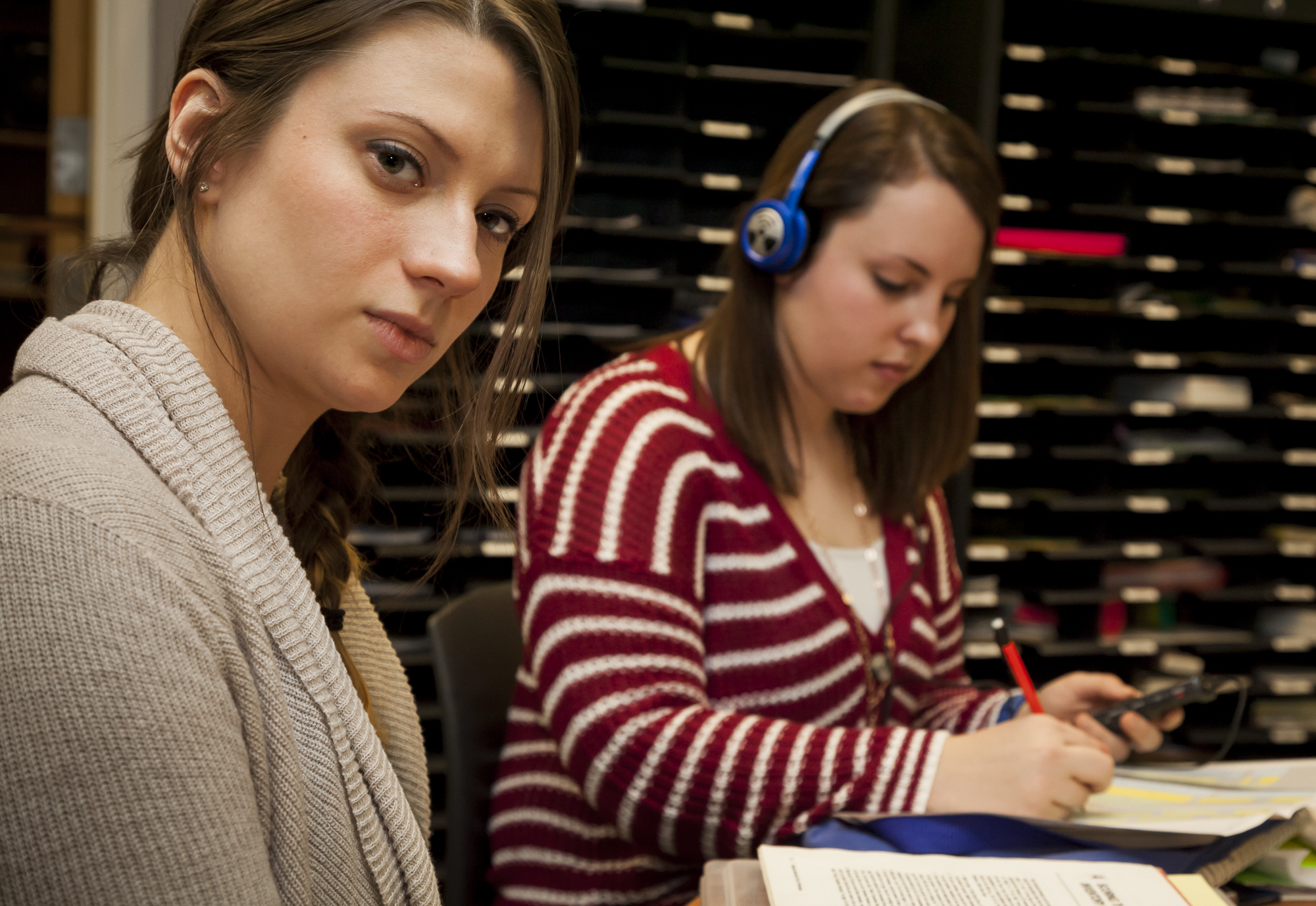 Two women sit at a table. One is looking at the camera and one is writing something on paper.