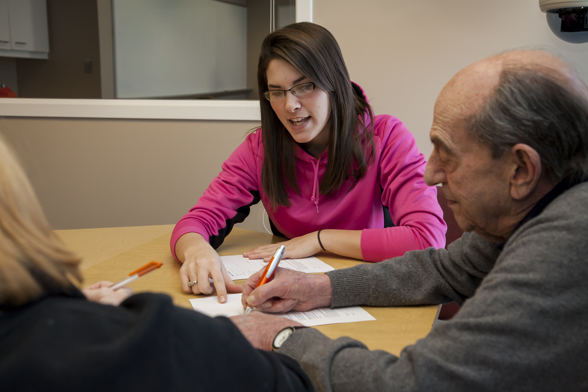 A graduate student clinician sits at a table with an elderly gentleman. She points at a form on the table, helping the man fill out the form. A women sits on the left hand side of the man, also filling out a form. 