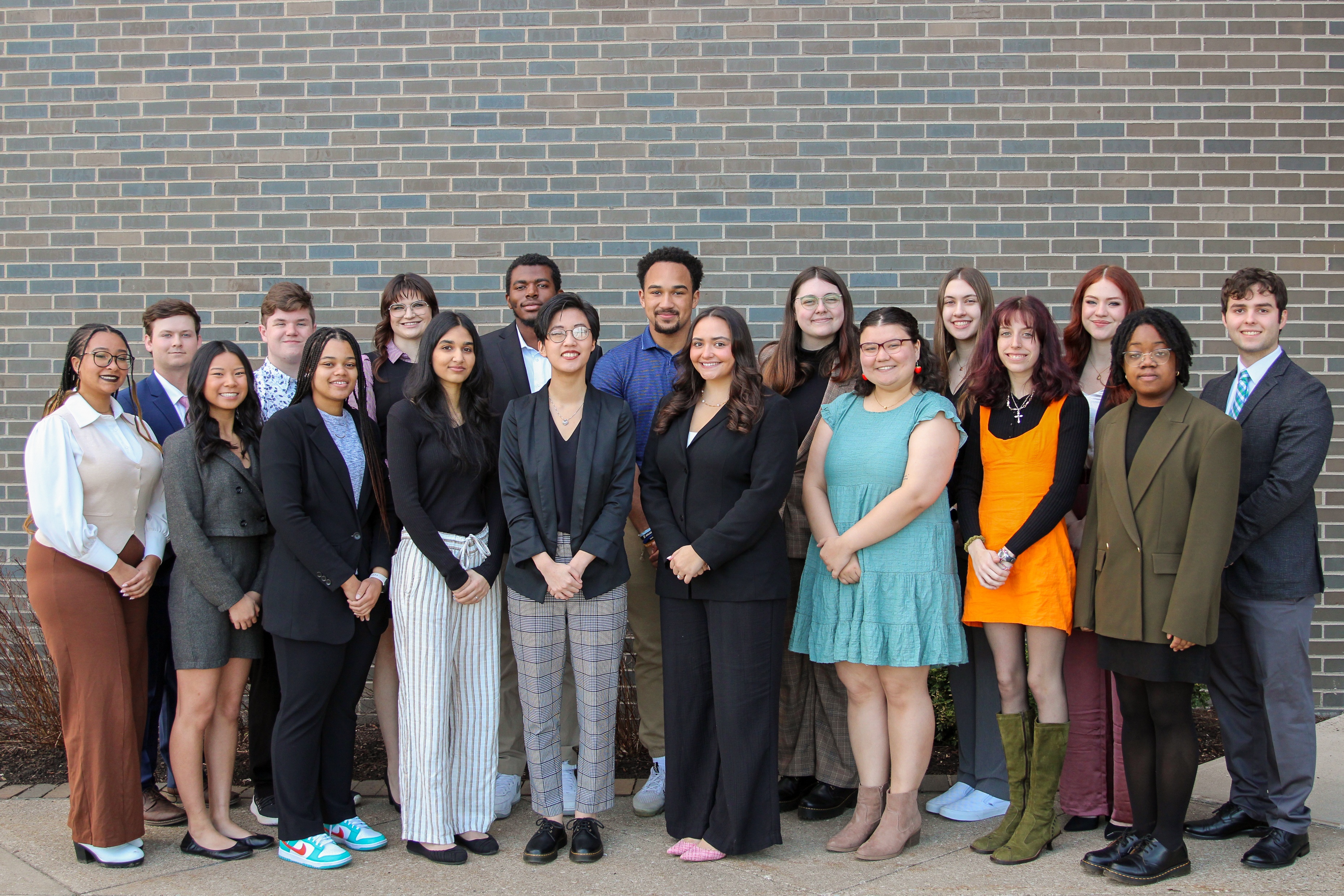 Group of 18 students dressed business professional in front of a multi-colored brick wall outside