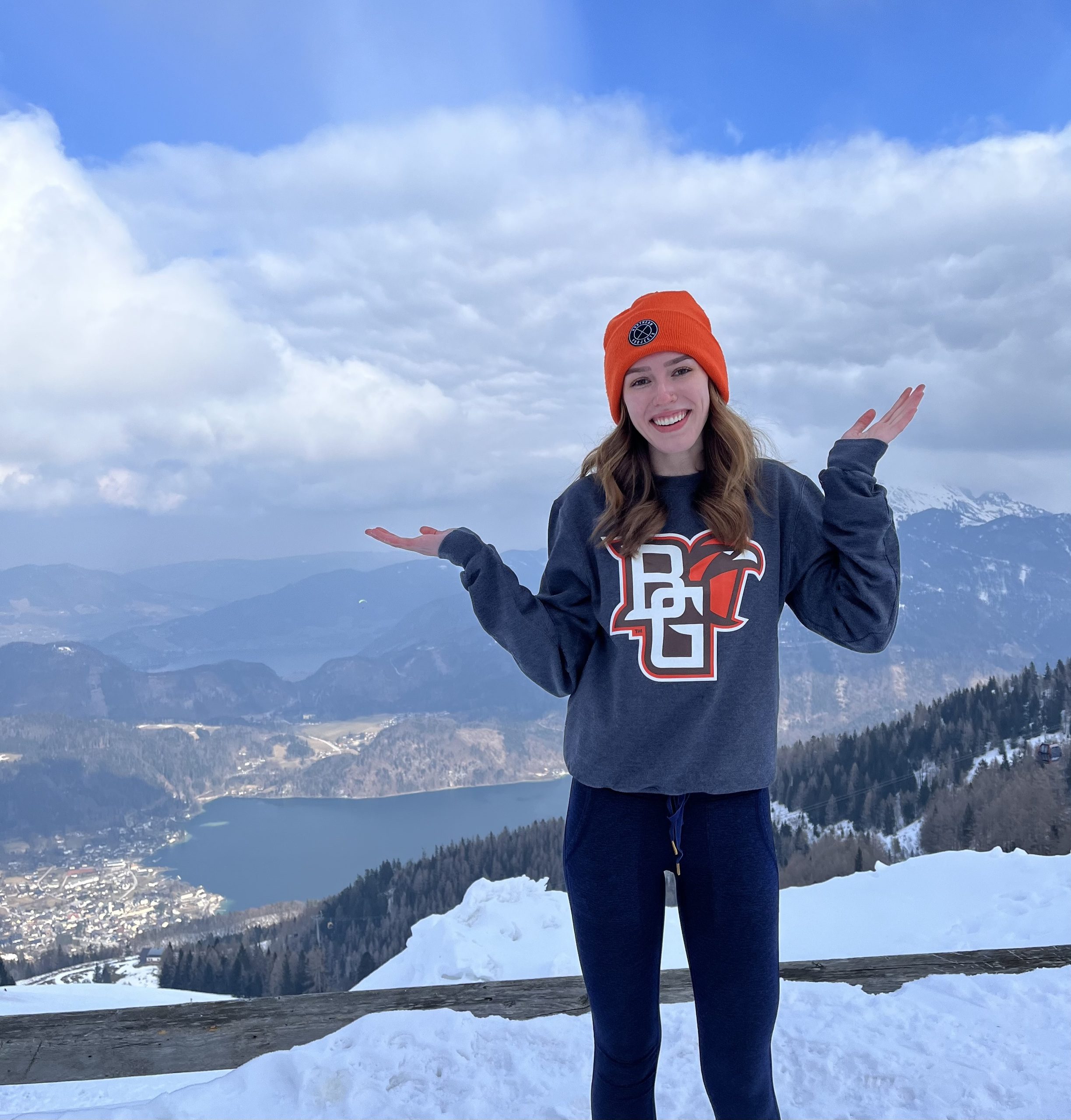 Student in BGSU sweatshirt and orange hat stands with hands out in front of a snowy mountain scenery