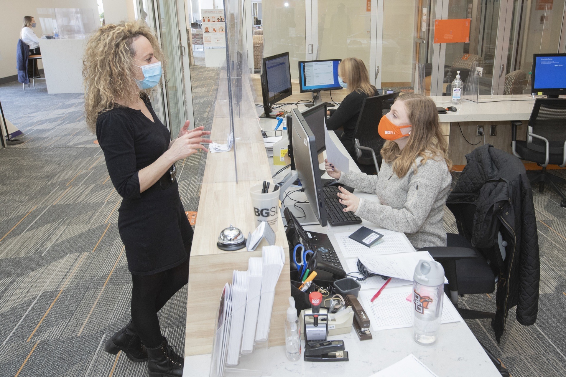 A female wearing a facemask speaks through plex-glass to a masked female employee at the BGSU Career Center.