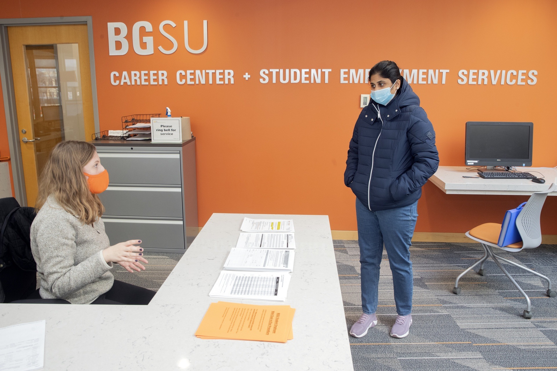 A female student wearing a facemask chats with a masked female employee of the BGSU Career Center at the front desk