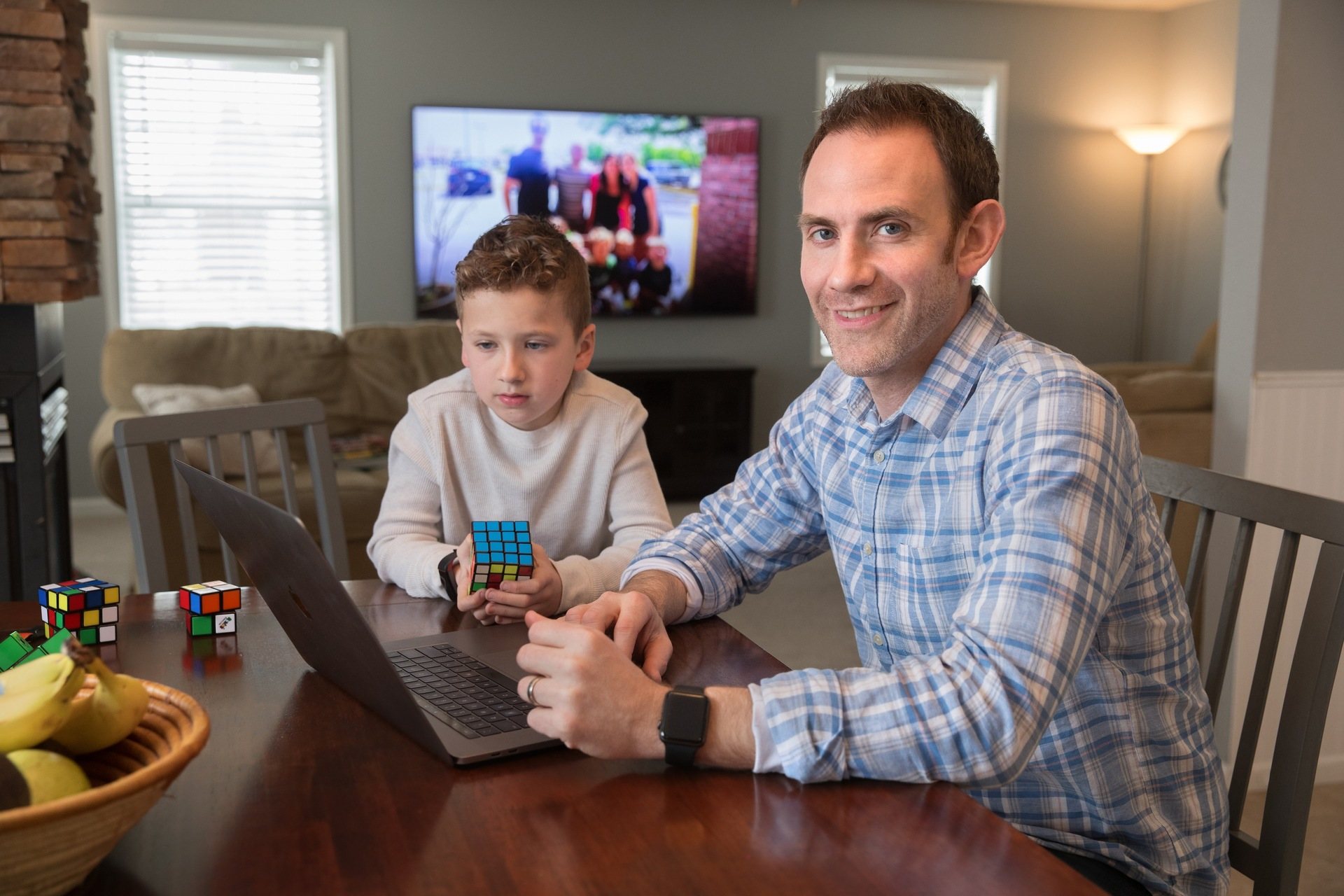 Man Smiling Working On Computer Next To Child Playing With Rubiks Cube.jpg