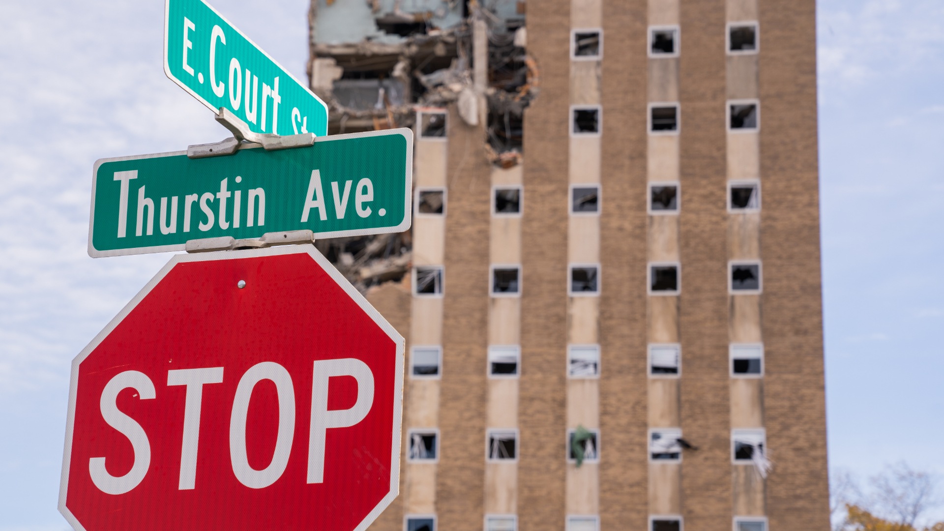 Administration building in the background with front wall missing on the top few floors and a stop sign with two street signs above it that read E. Court St. and Thurstin Ave.