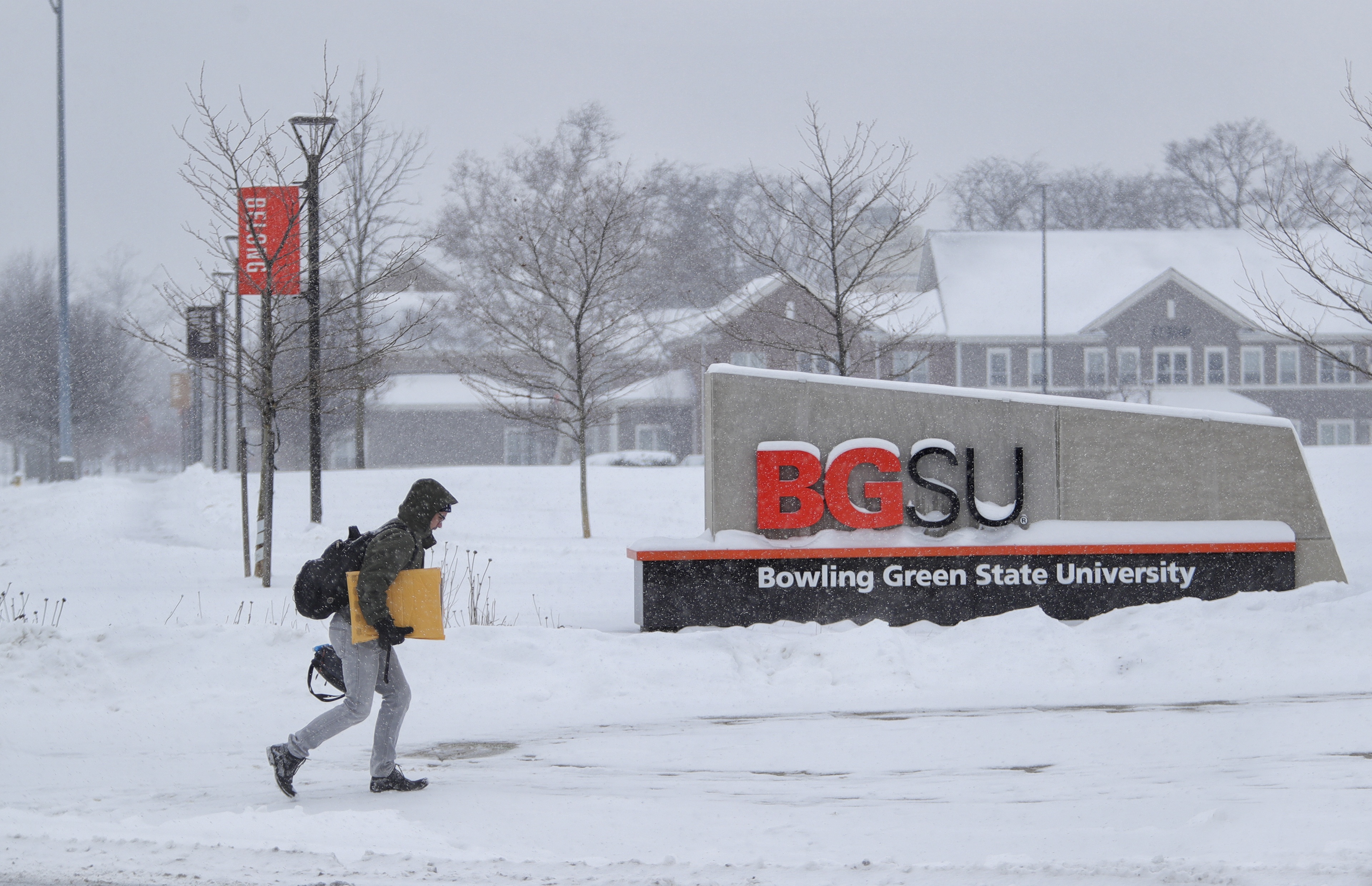 A student walks in front of the BGSU sign by Greek Village heading to campus with their arms full of materials for a class. 