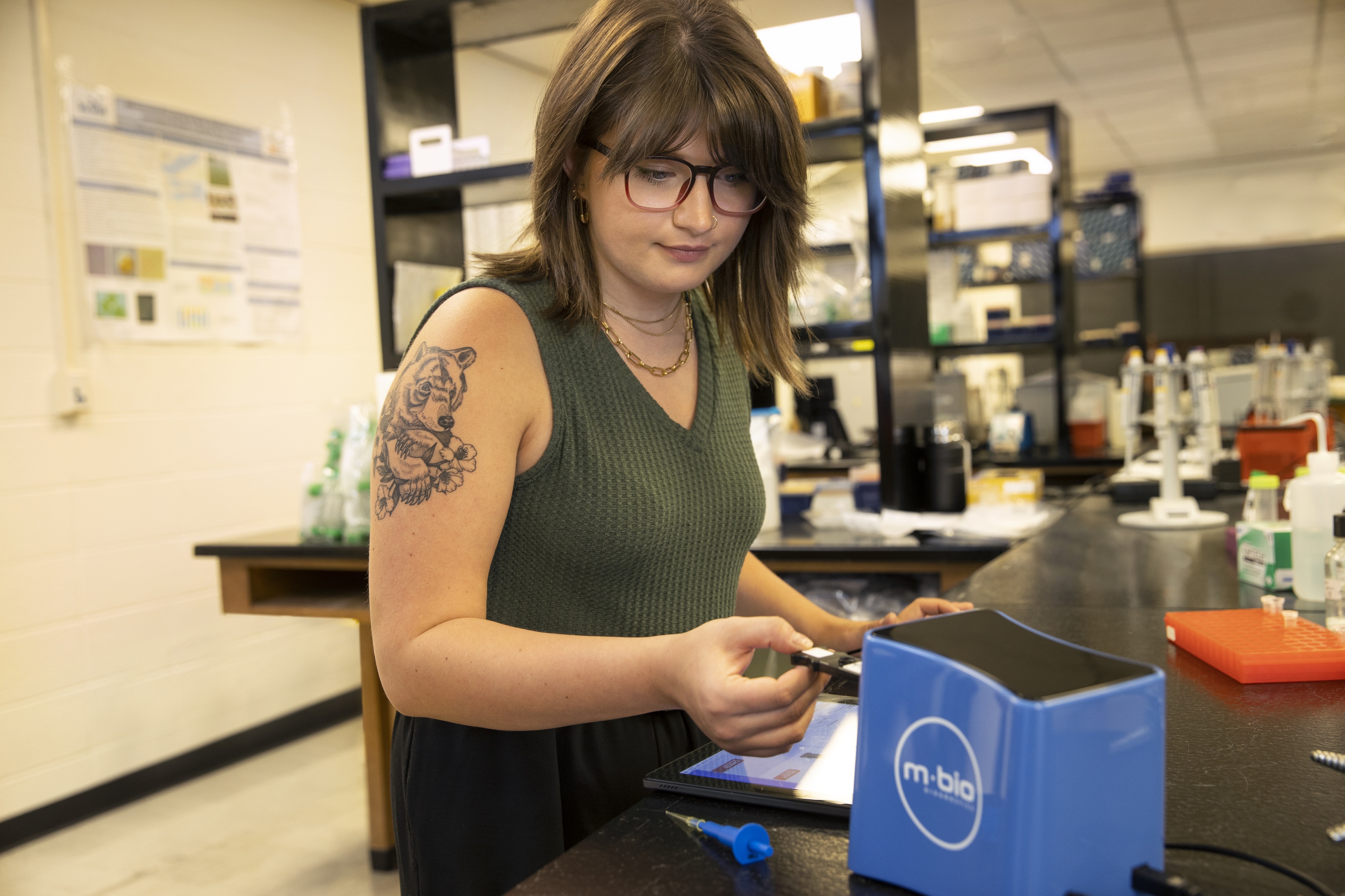 student, Katie Barker, standing at a counter with an ipad, holding a piece of equipment