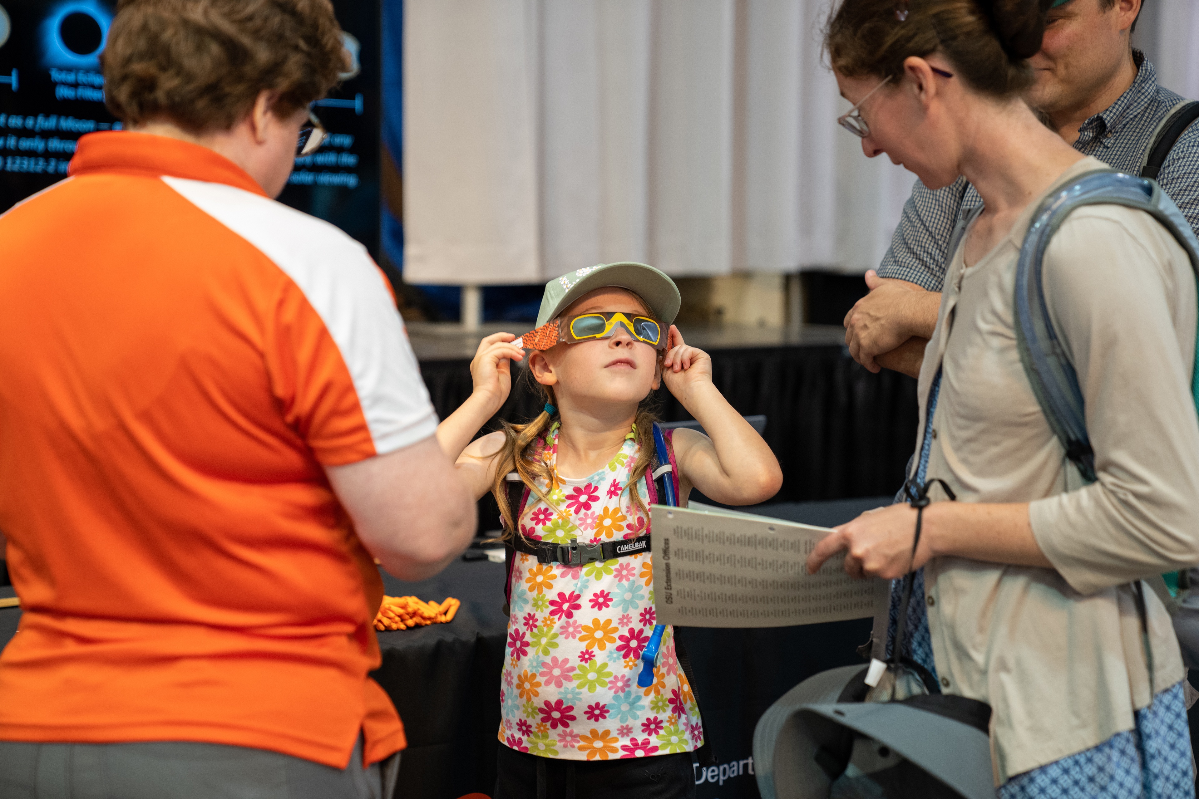 Two adults watch a girl put on brown and orange eclipse glasses