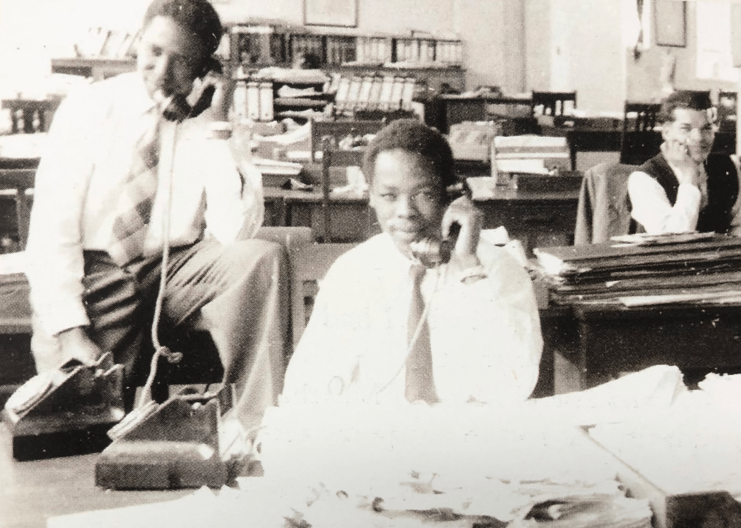 Two young men hold rotary phone handsets to their ears while another young man in the background watches