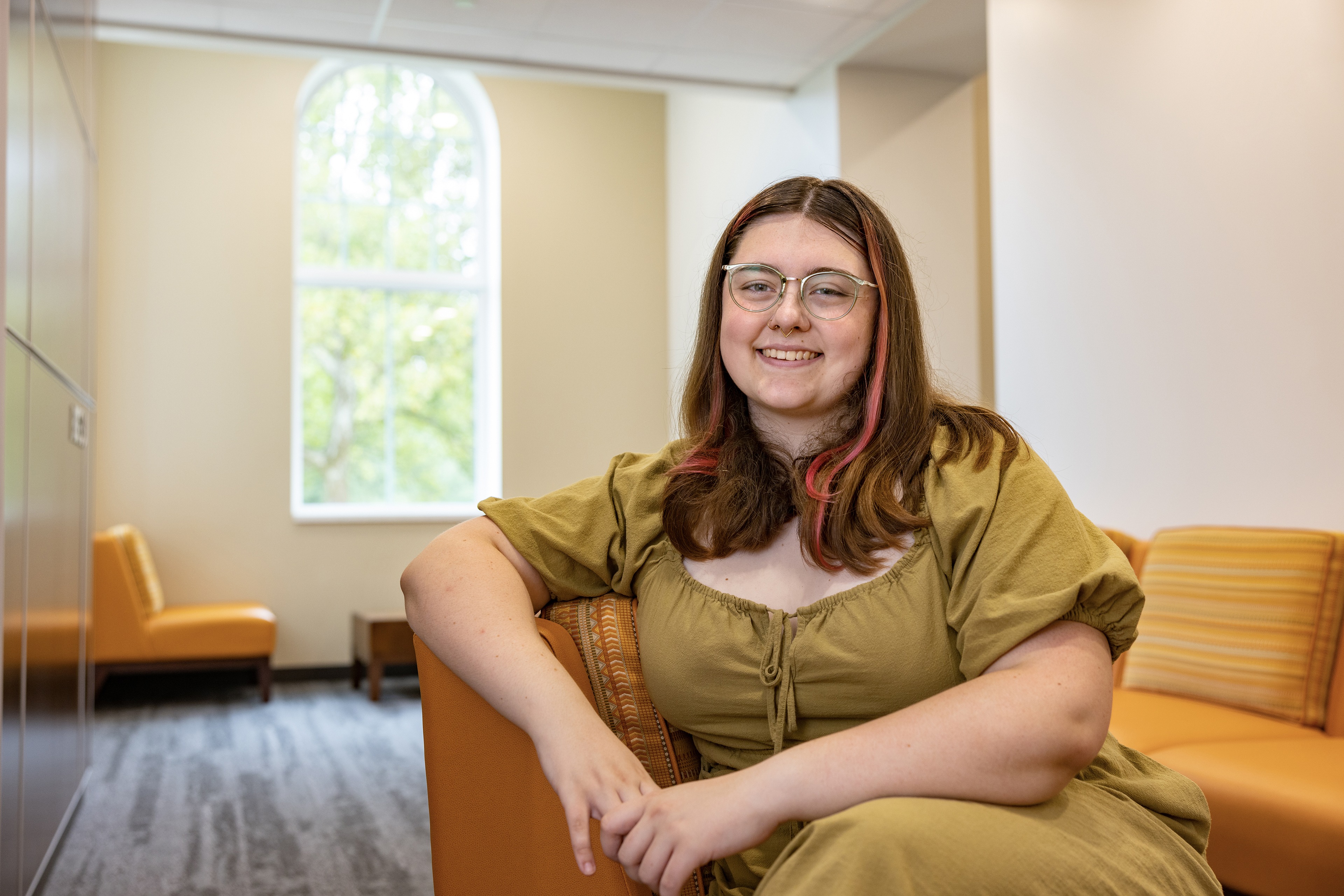 Student Elyse is in a green dress and sits with their arm around the back of an orange chair