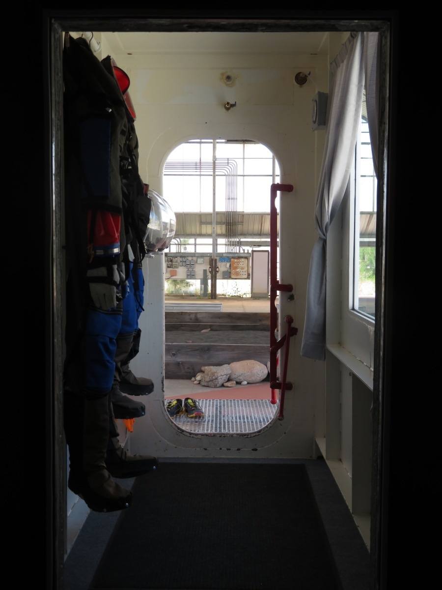 An interior close-up of a narrow hallway in the Space Analog for the Moon and Mars in Oracle, Arizona. The module is made of white metal and has a red stripe down the side. The windows are dark and there are no astronauts visible. Spacesuits hang from the wall on the left and an interior door is open straight ahead.