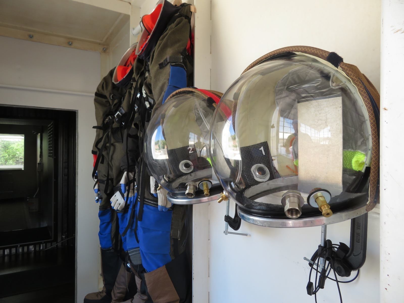 Close-up image of two helmets and space suits hanging on the wall in a room at the Space Analog for the Moon and Mars. The helmets are white with black visors. The space suits are orange and blue. The helmets are hanging on the left side of the wall, and the space suits are hanging on the right side of the wall.