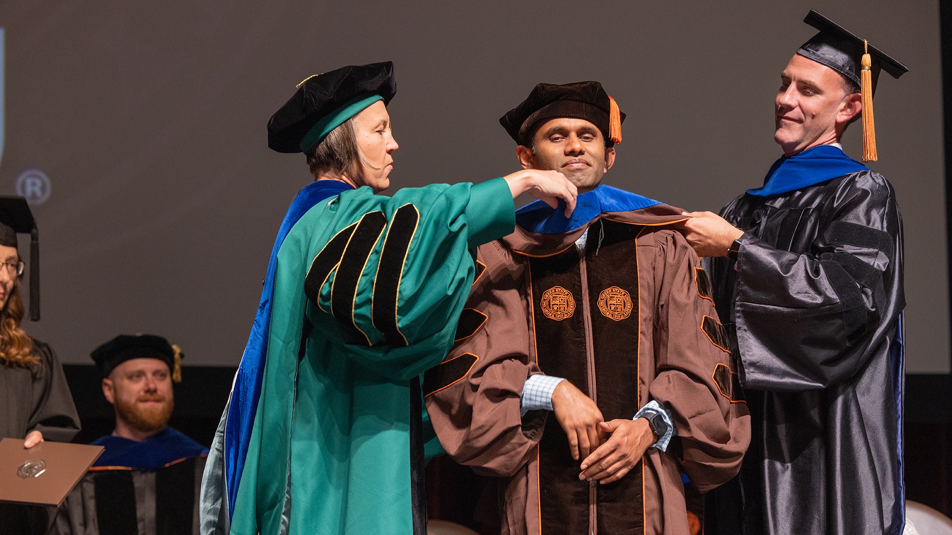 Three people wearing graduation gowns at the BGSU Hooding Ceremony for doctoral graduates.