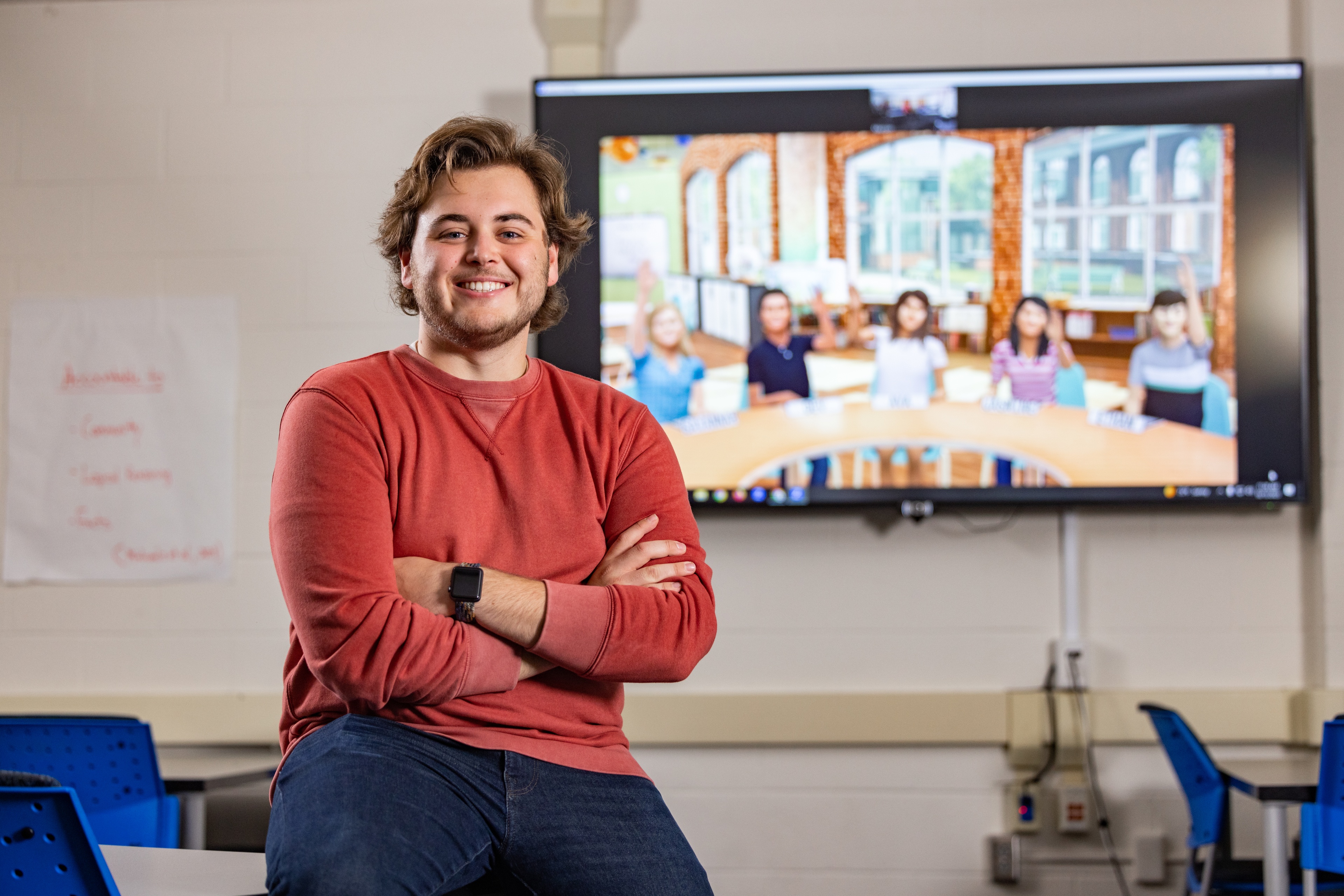 BGSU student smiles at the camera with a screen showing five simulated students in the Mursion display in the background.