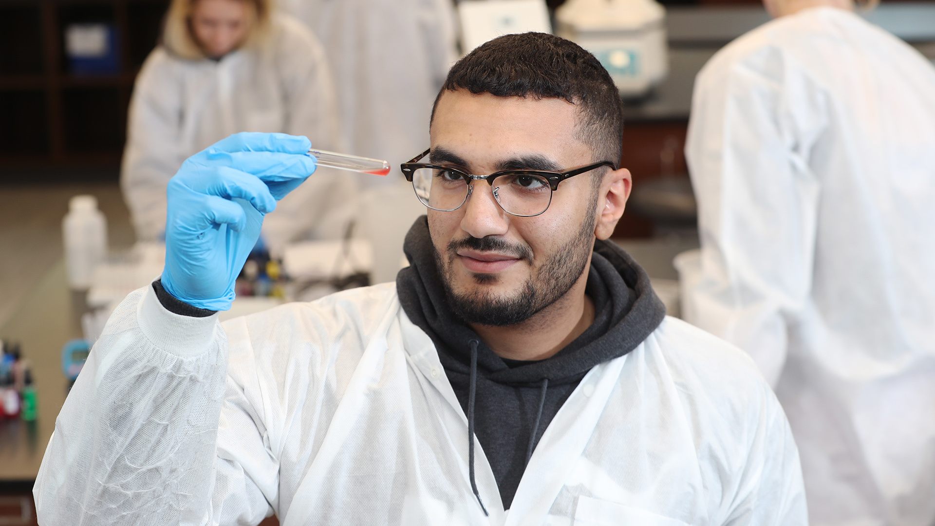 A person wearing glasses, gloves and a white lab coat holding a test tube with red liquid. 