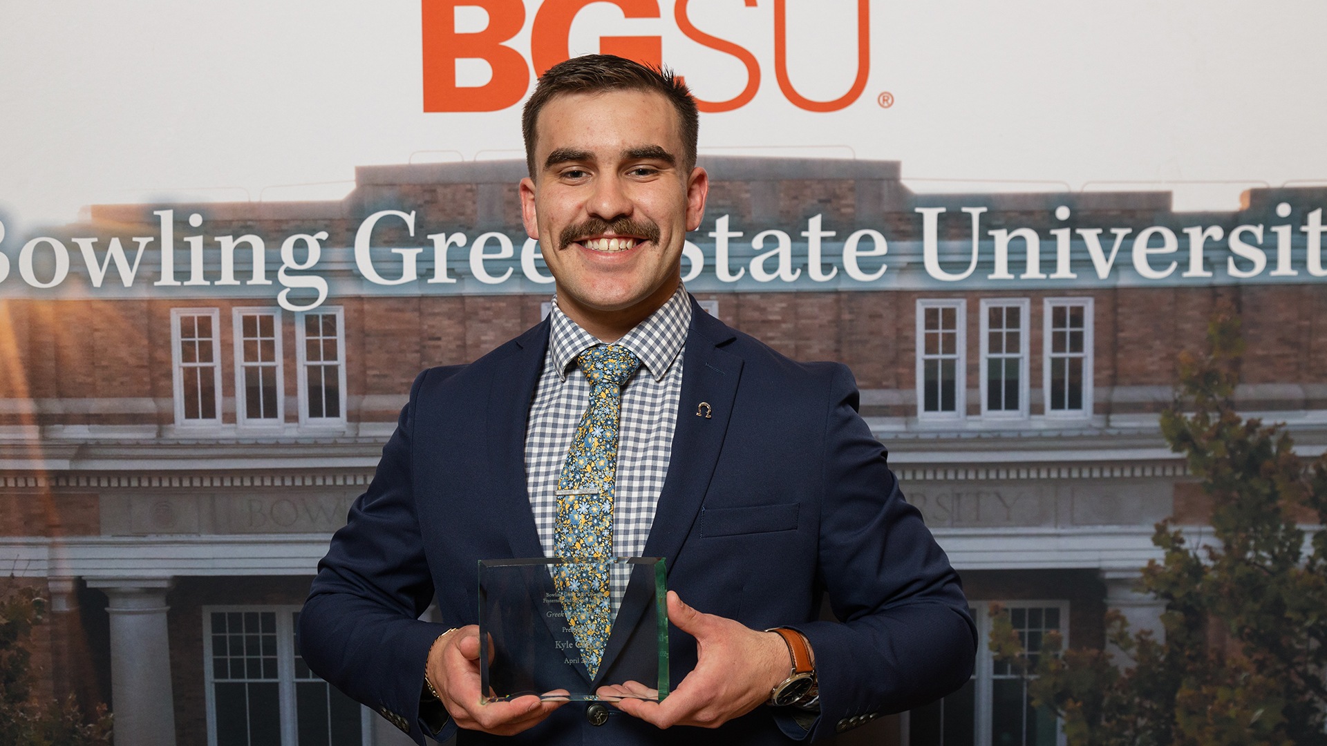 Kyle Calvey holds a glass trophy in front of a BGSU mural.