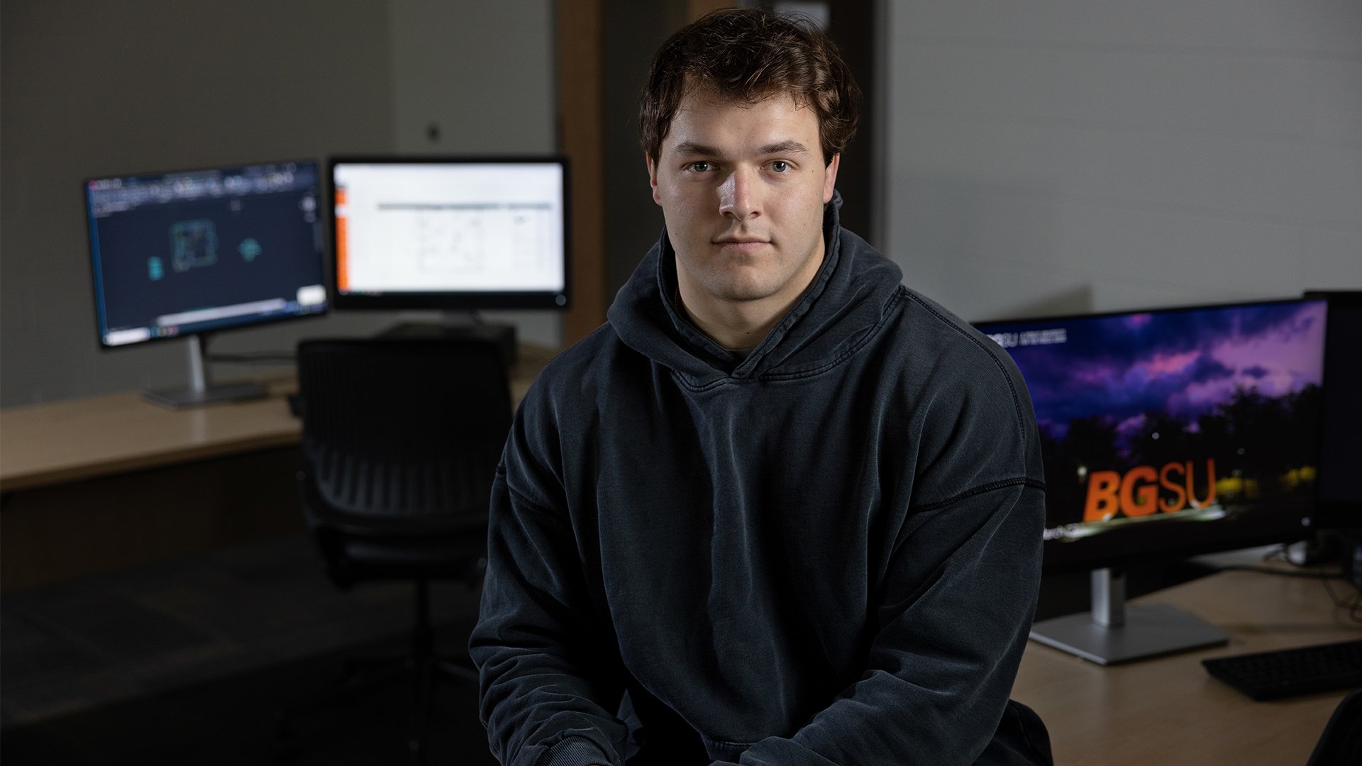 A person sitting on a desk with computers in the background.