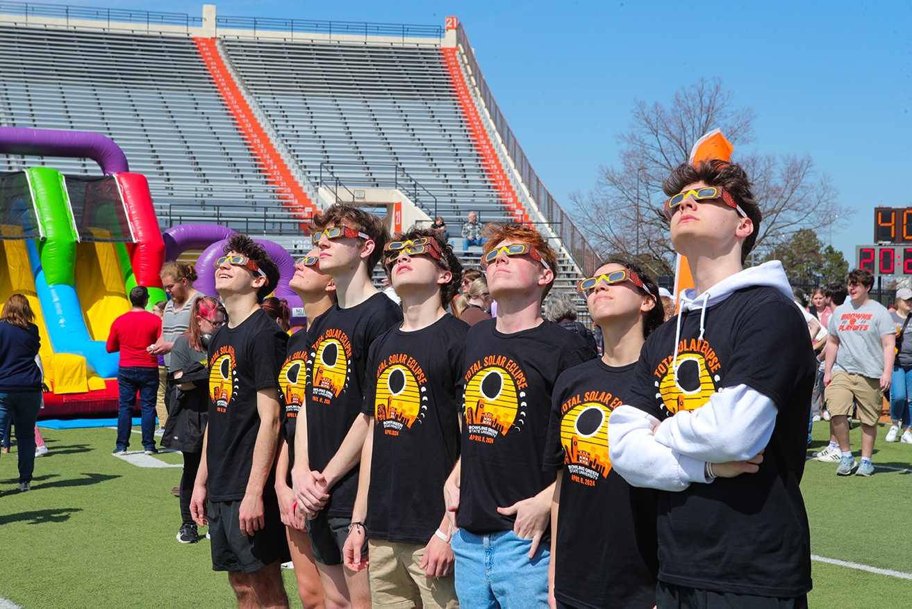 Seven students wearing matching T-shirts and eclipse glasses stand in a row