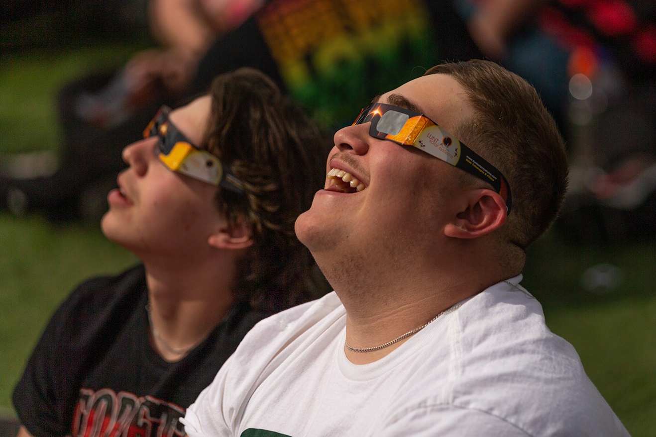 Two people wear eclipse glasses and look up 