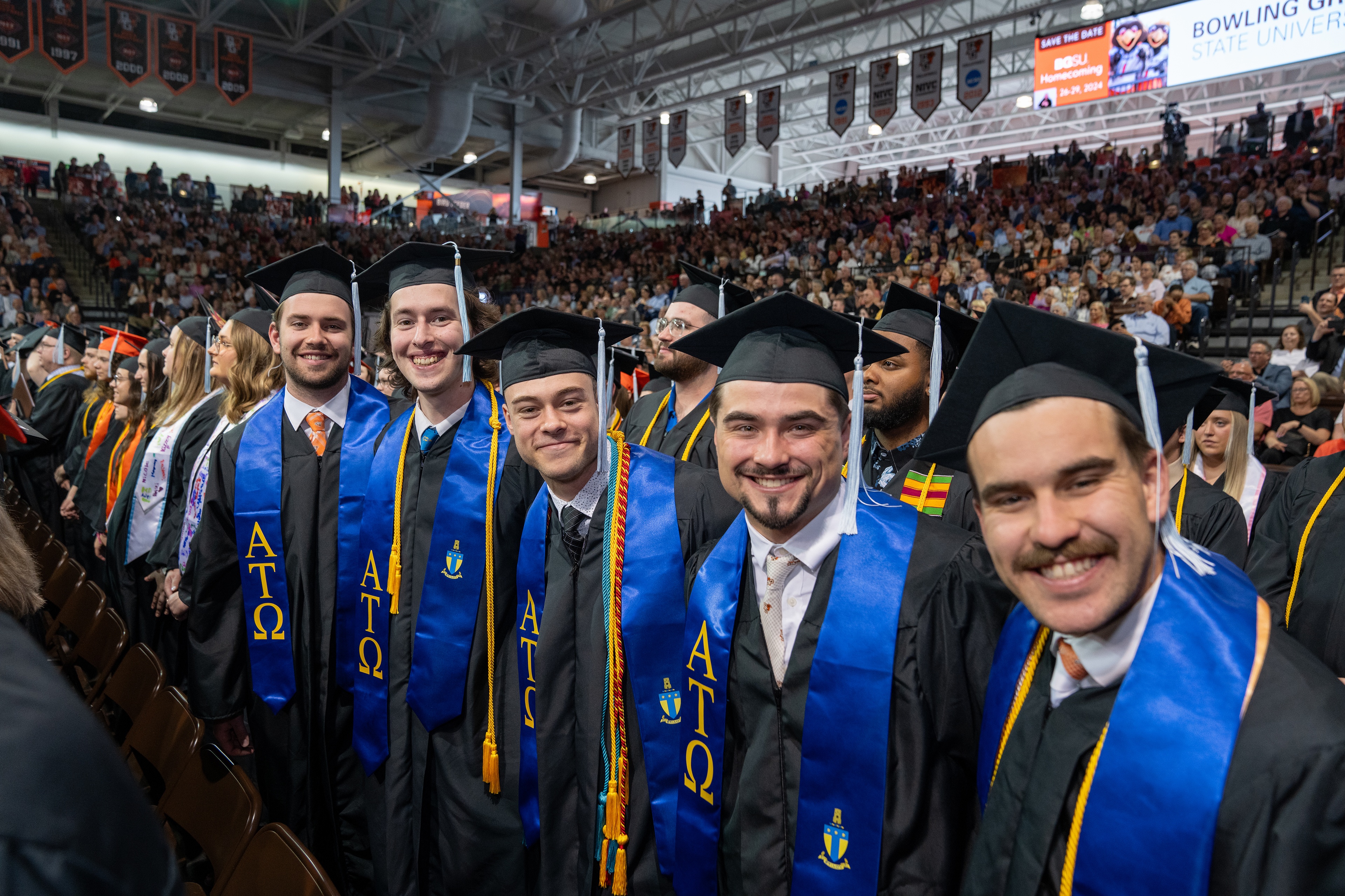 Five young men in graduation gowns with sashes stand together at graduation