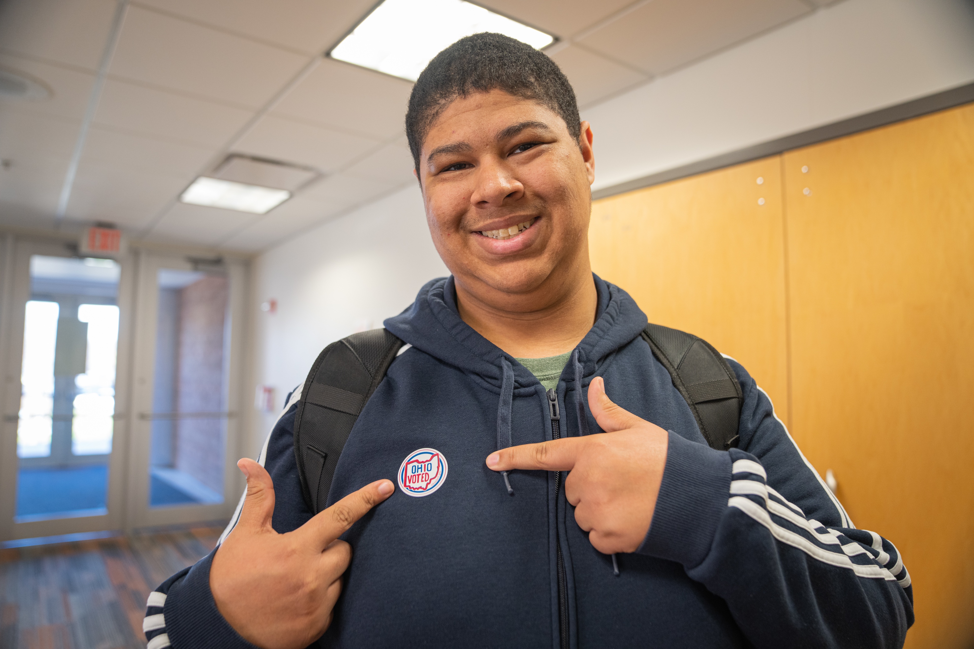 A person smiles and points to a voting sticker on their sweatshirt.