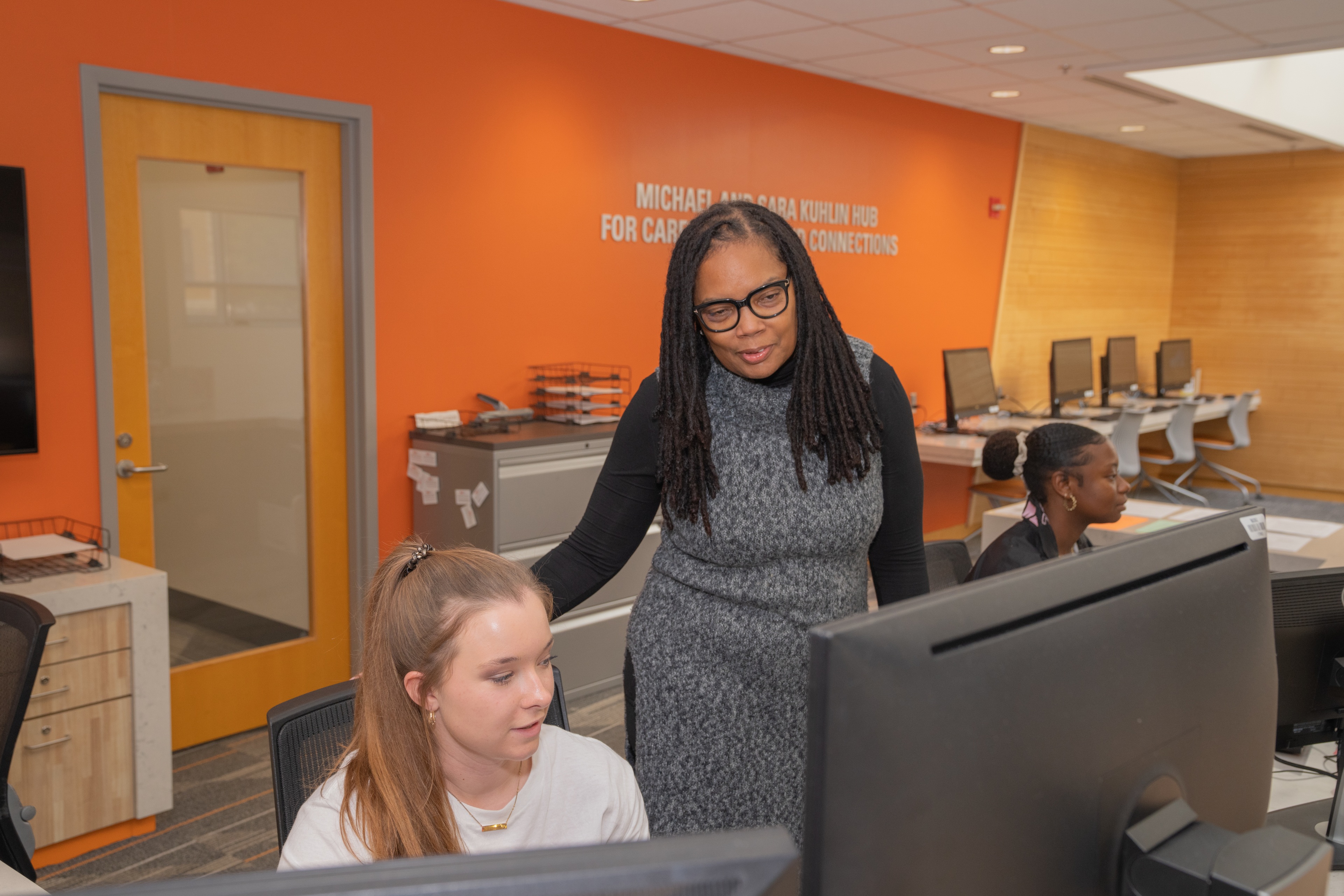 A person stands and looks at a seated student working on a computer 