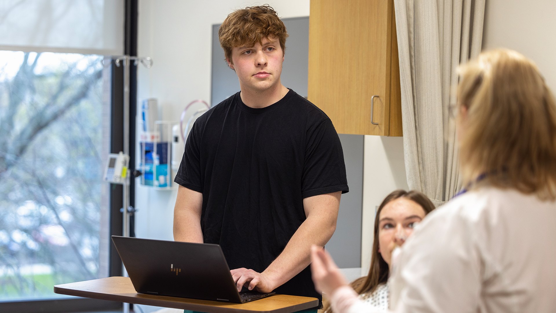 A person types into a laptop while standing next to a patient's bed.