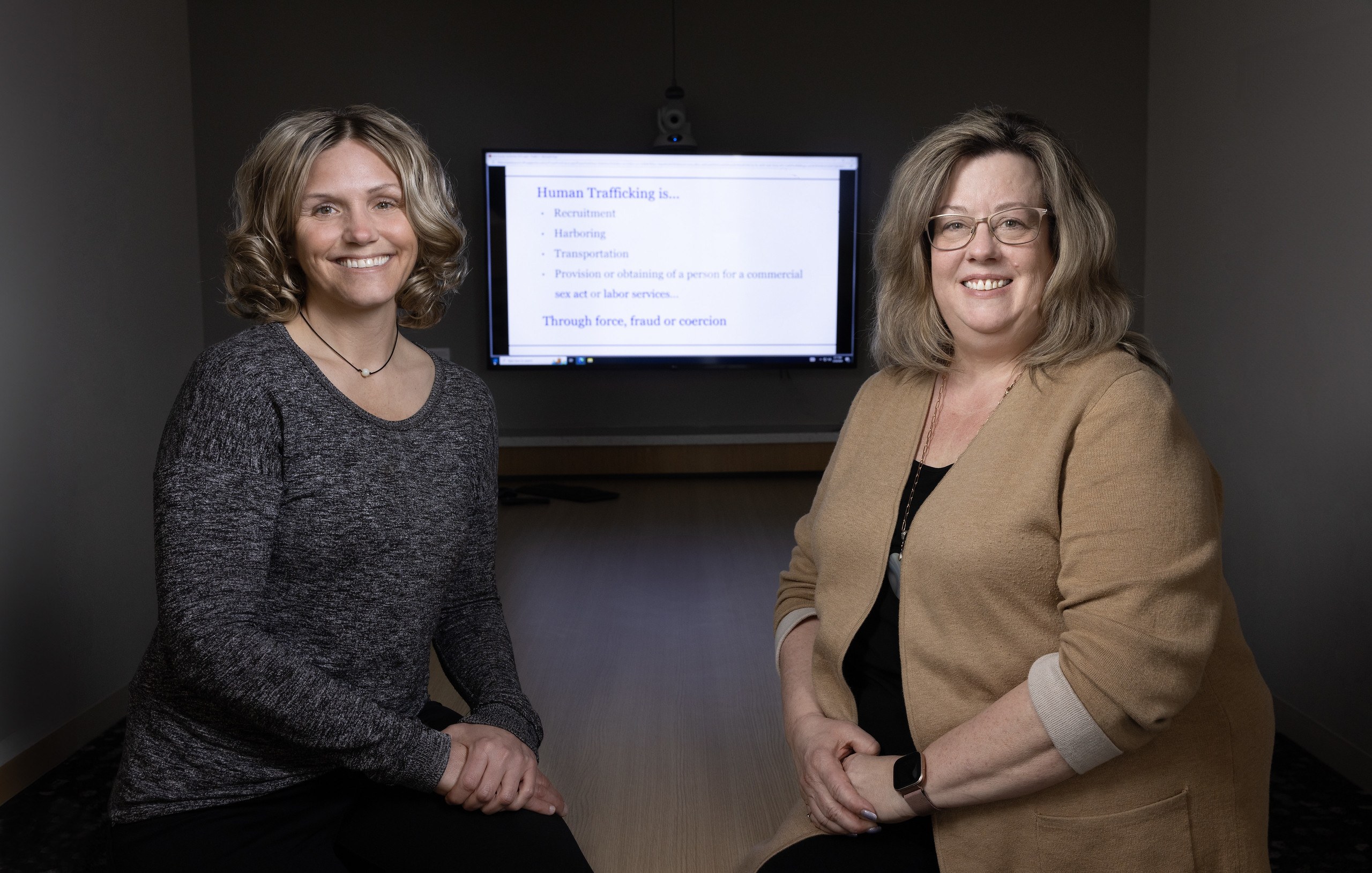 Two women smile at the camera with a presentation on human trafficking in the background.