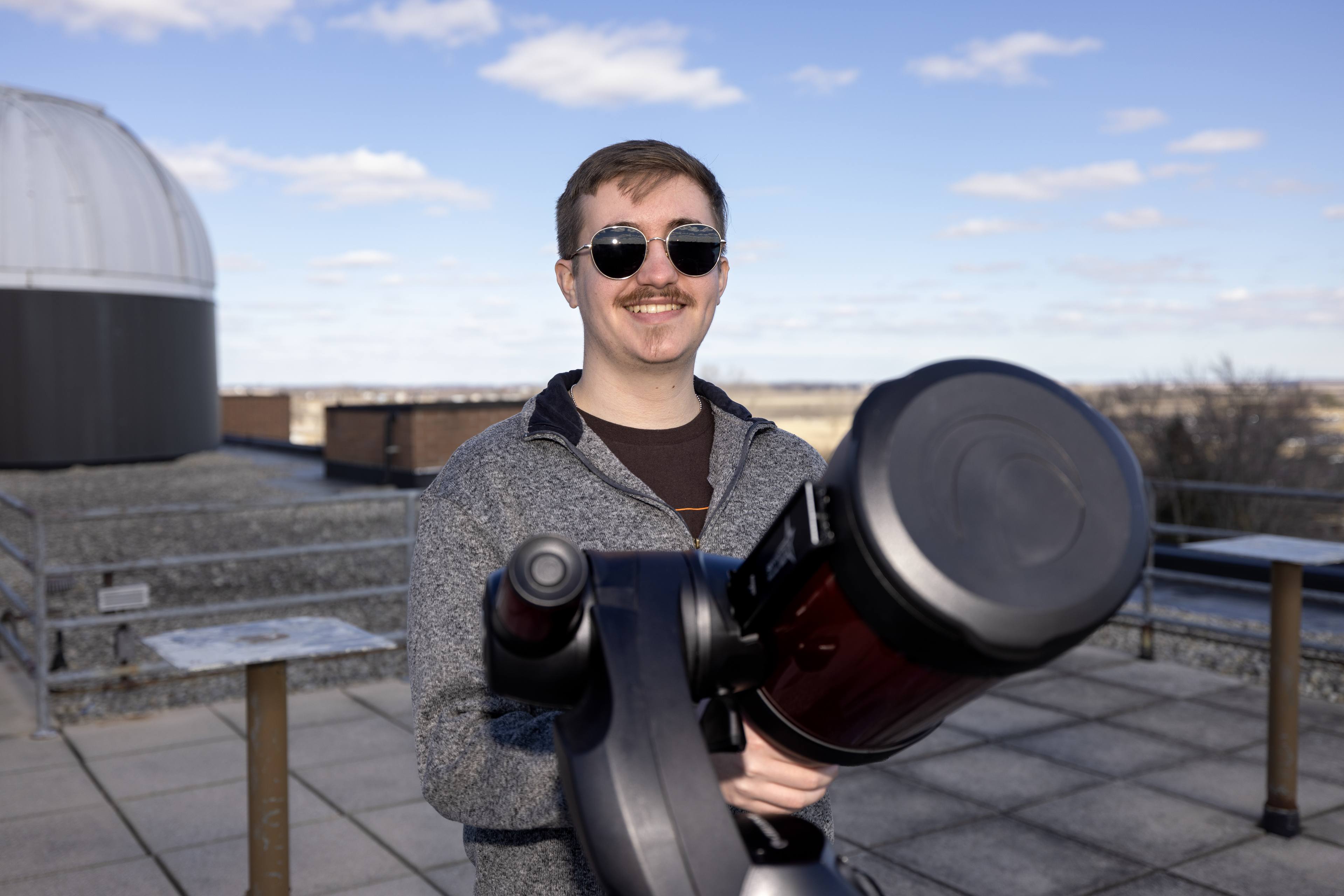 A student smiles as he stands behind a telescope.