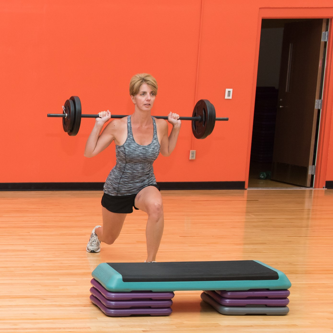 Woman performing a lunge while holding a barbell over her shoulders, wearing a gray tank top and black shorts. 