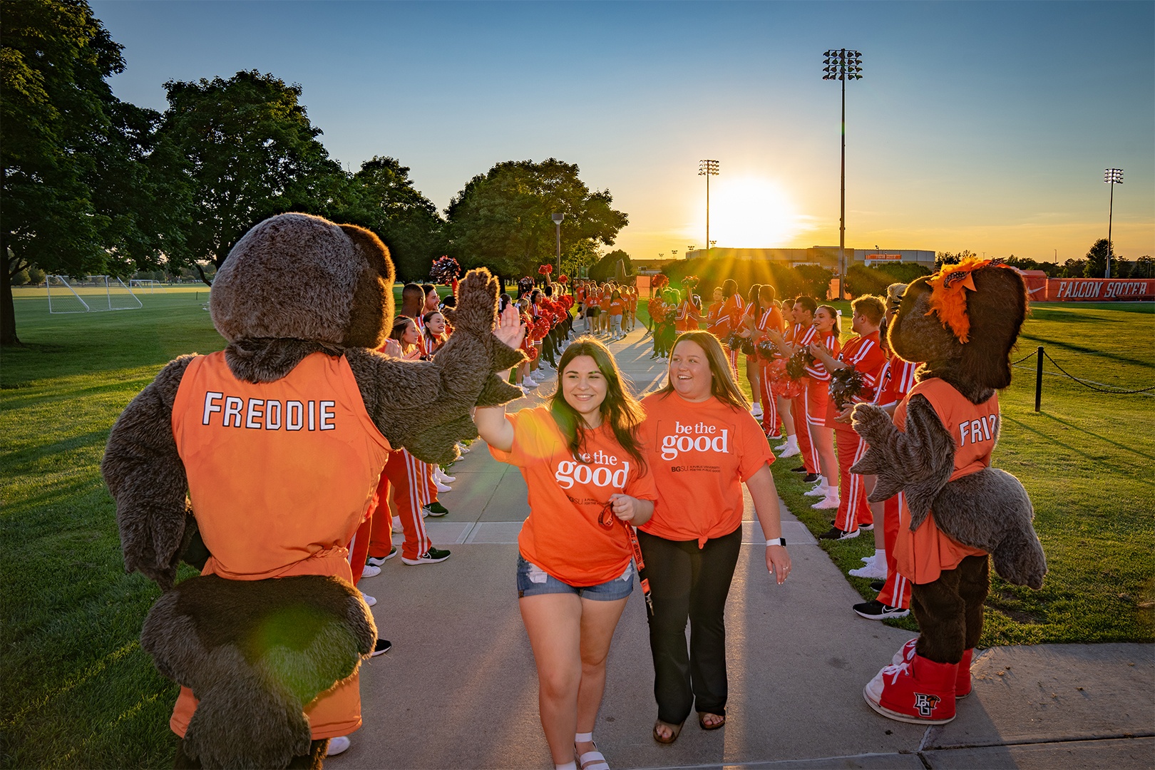 Two students walking down sidewalk, giving Freddie Falcon a high-five