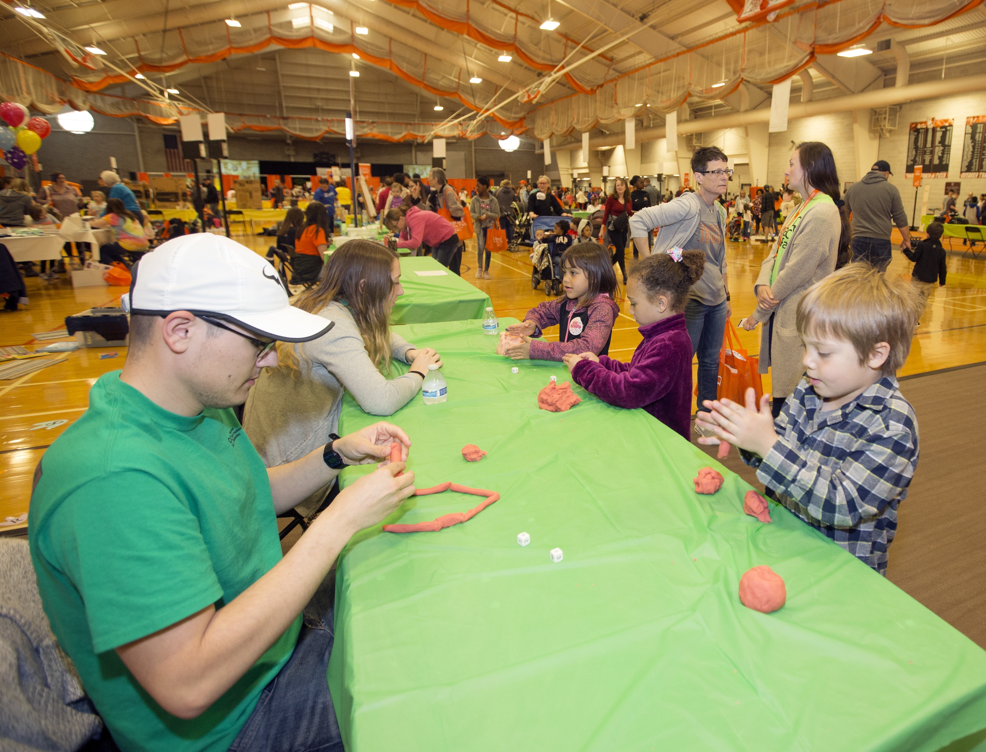 A male art education master’s student from BGSU and a little boy play with clay at a community art education event with many tables, exhibits and other students and children.