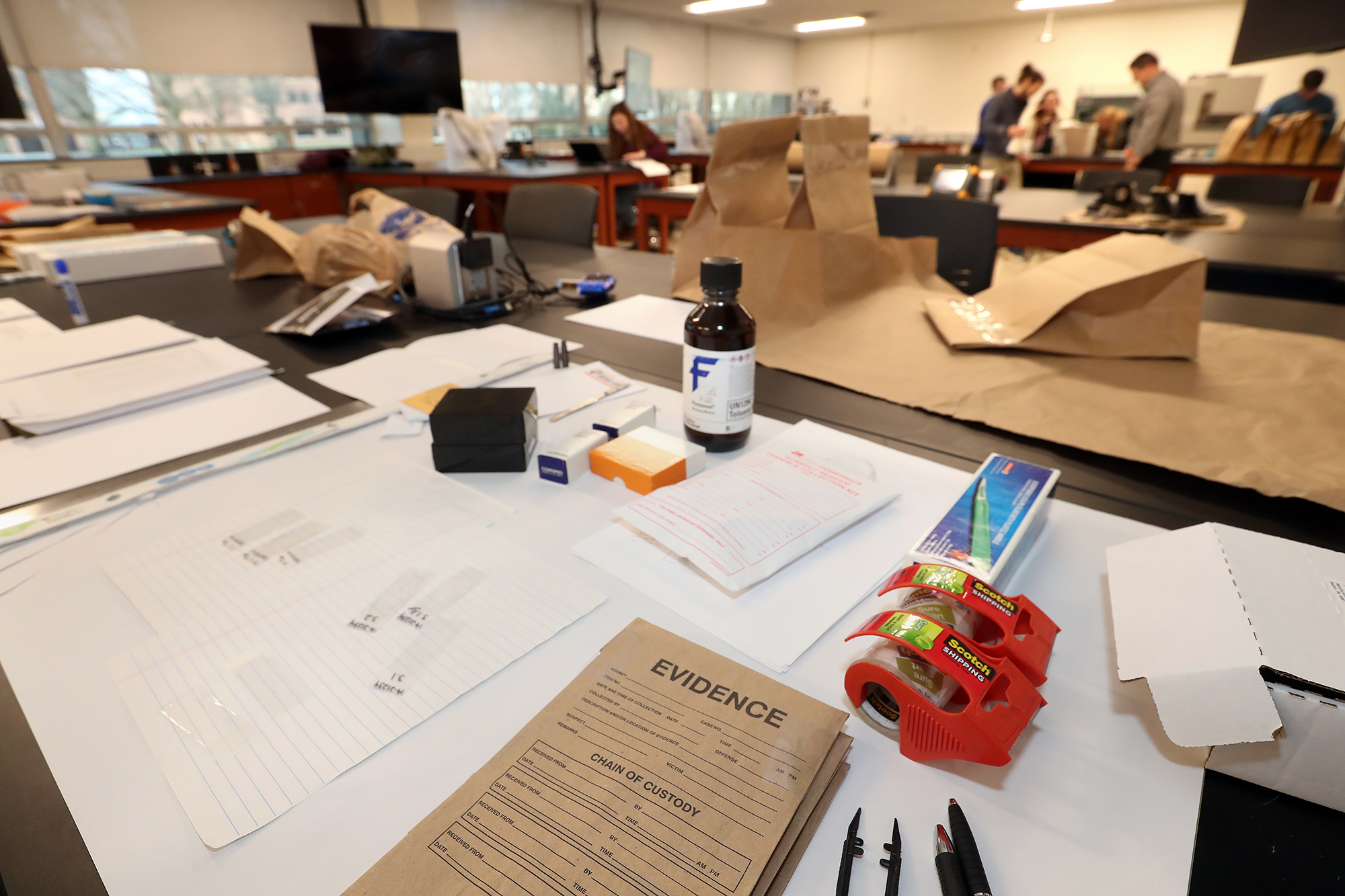 A table in the BGSU forensic lab features brown paper evidence bags, chemicals and others tools that the students in the background use to process crime scene evidence.