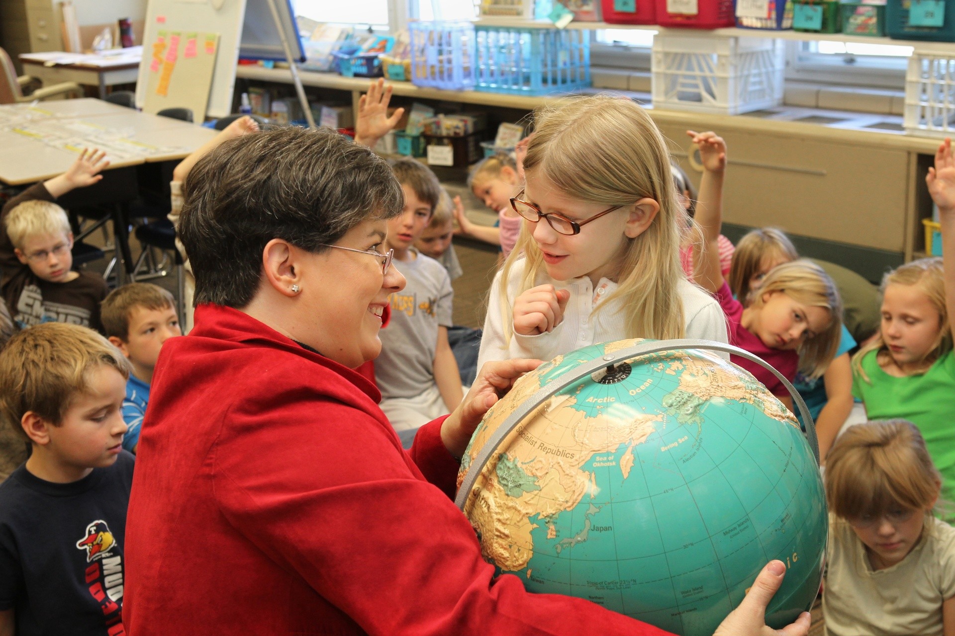 A female world language teacher points out a country on a globe to a little girl as the rest of the class sits on the floor around them.