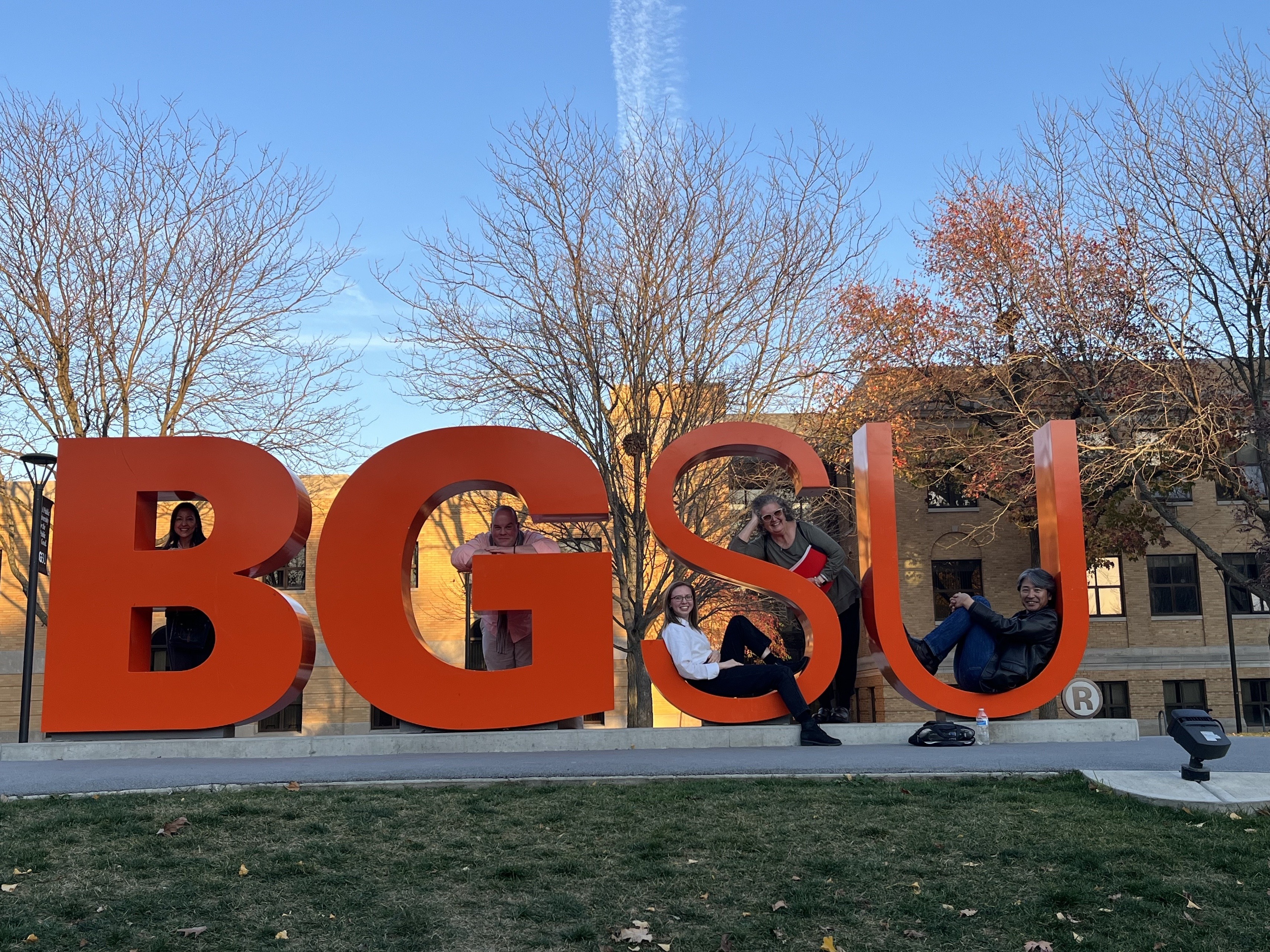 A photo of Paul Kei Matsuda and English Department faculty posing with the giant BGSU letters. Left to right: Fernanda Capraro faces camera through the top of letter B, Chad van Buskirk leans on letter G, Anastasiia Kryzhanivska and Lucinda Hunter are inside letter S, and Paul Kei Matsuda is sitting inside letter U. 