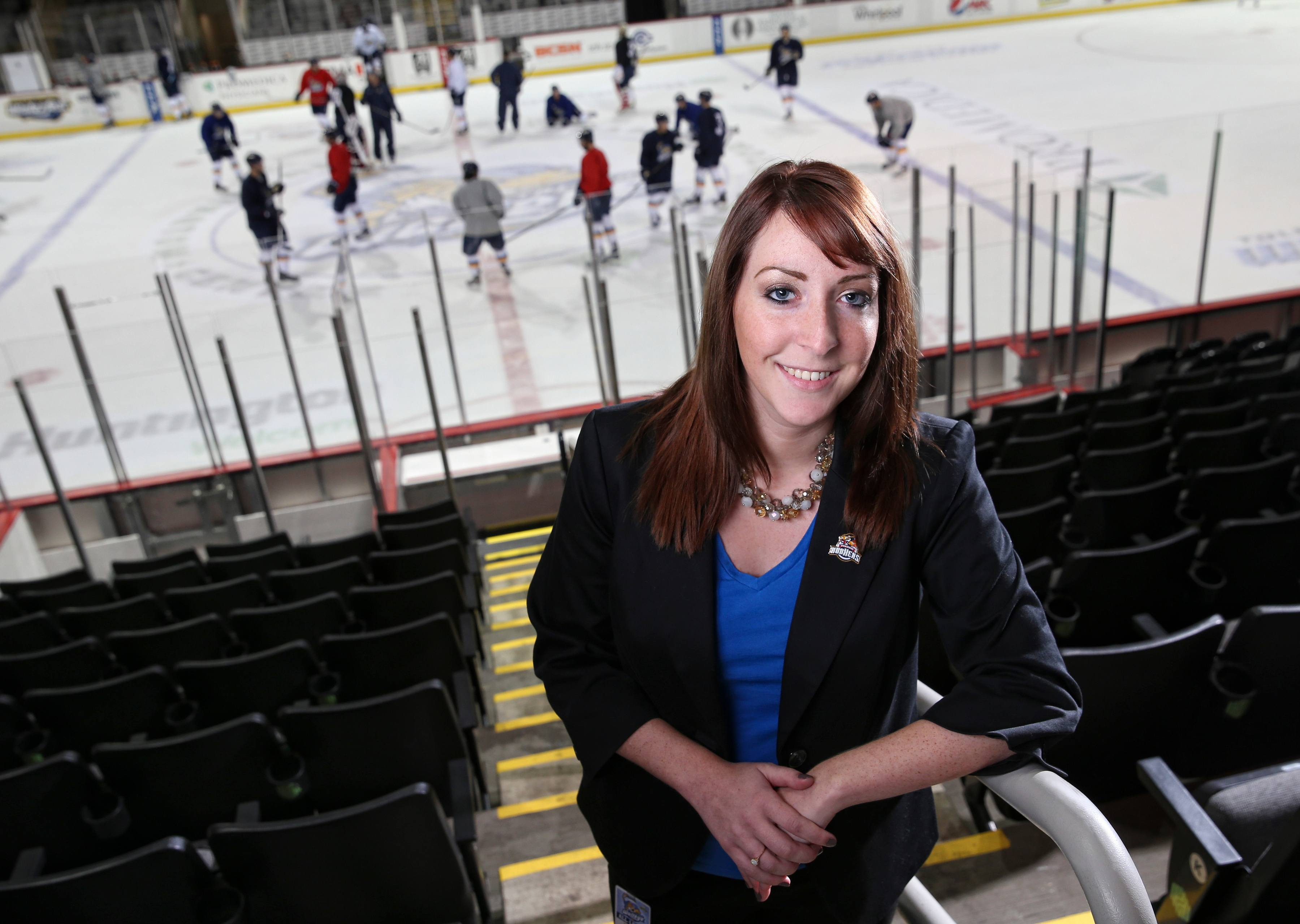 A woman in the BGSU sport administration master's program stands in the bleachers of a hockey rink as players practice on the rink behind her.