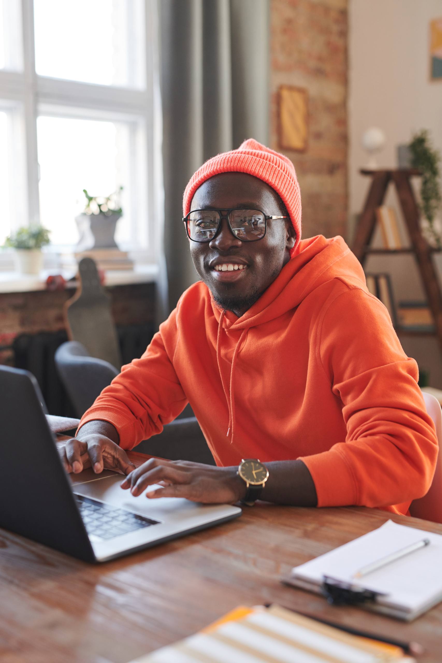 Vertical portrait of joyful African American guy wearing bright orange outfit sitting at desk at home surfing Internet on laptop smiling at camera