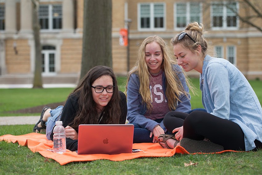 Three students playing in grass in front of BGSU's Bowen-Thompson Student Union looking at a laptop