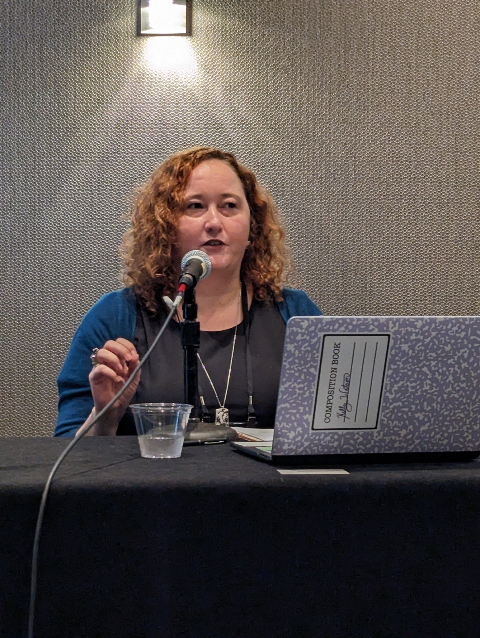  White woman with curly red hair is seated at a table, delivering presentation to audience