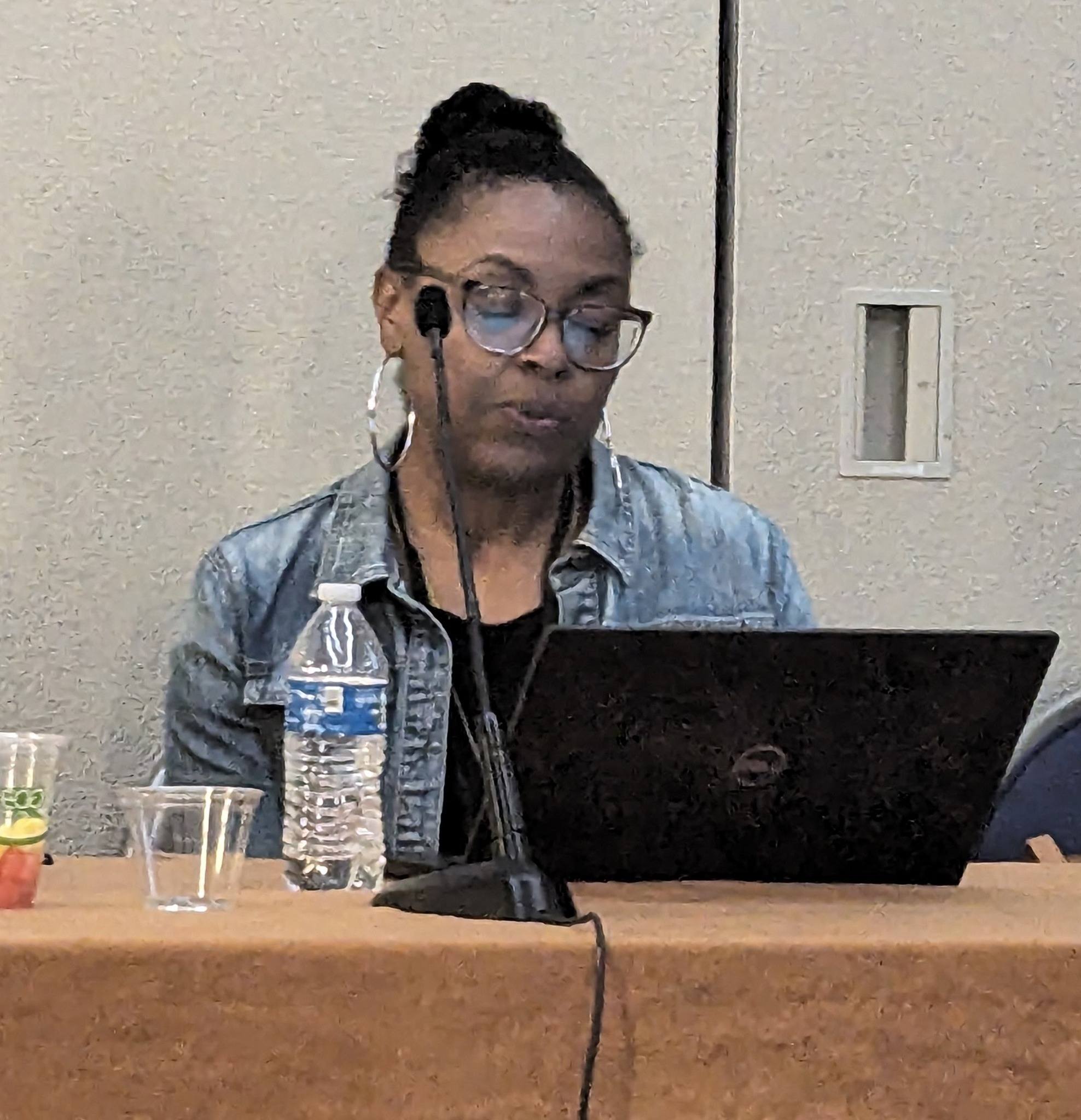 Black woman with glasses and hair pulled back is seated at a table, delivering presentation to audience
