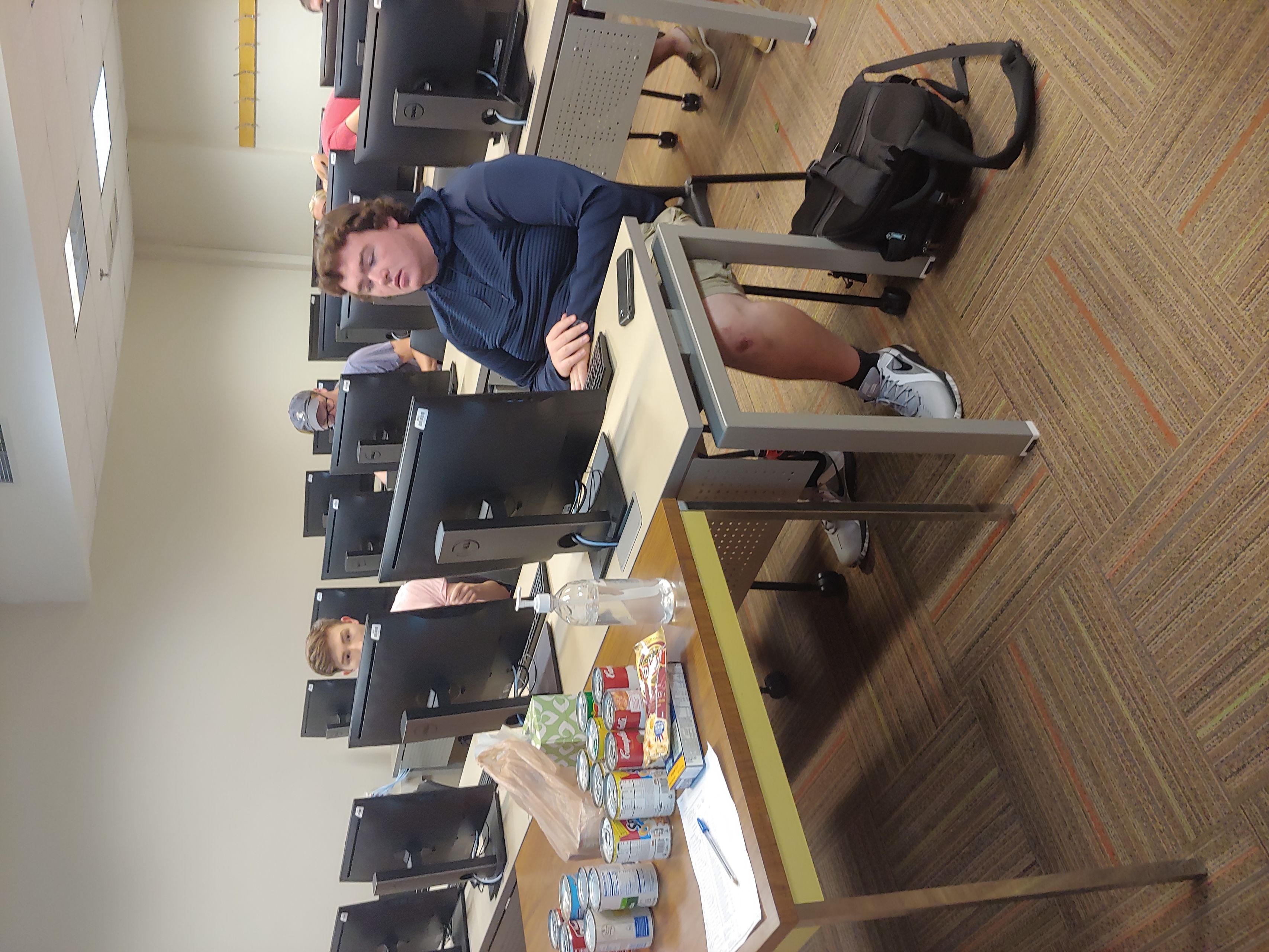 Students sit at computers; on the tables in front of them are cans and boxes of food