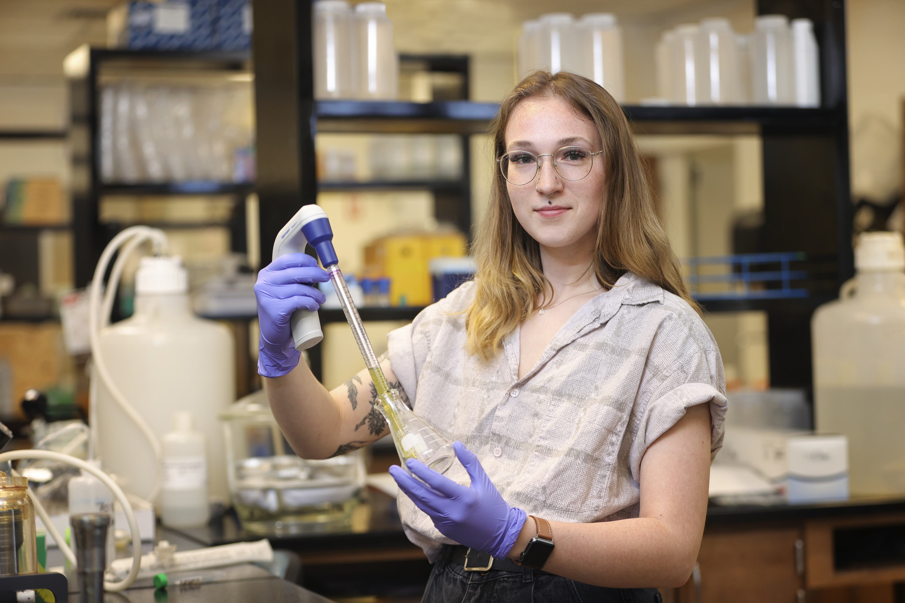student, Kate Brown, holding a beaker and other testing equipment while wearing purple gloves